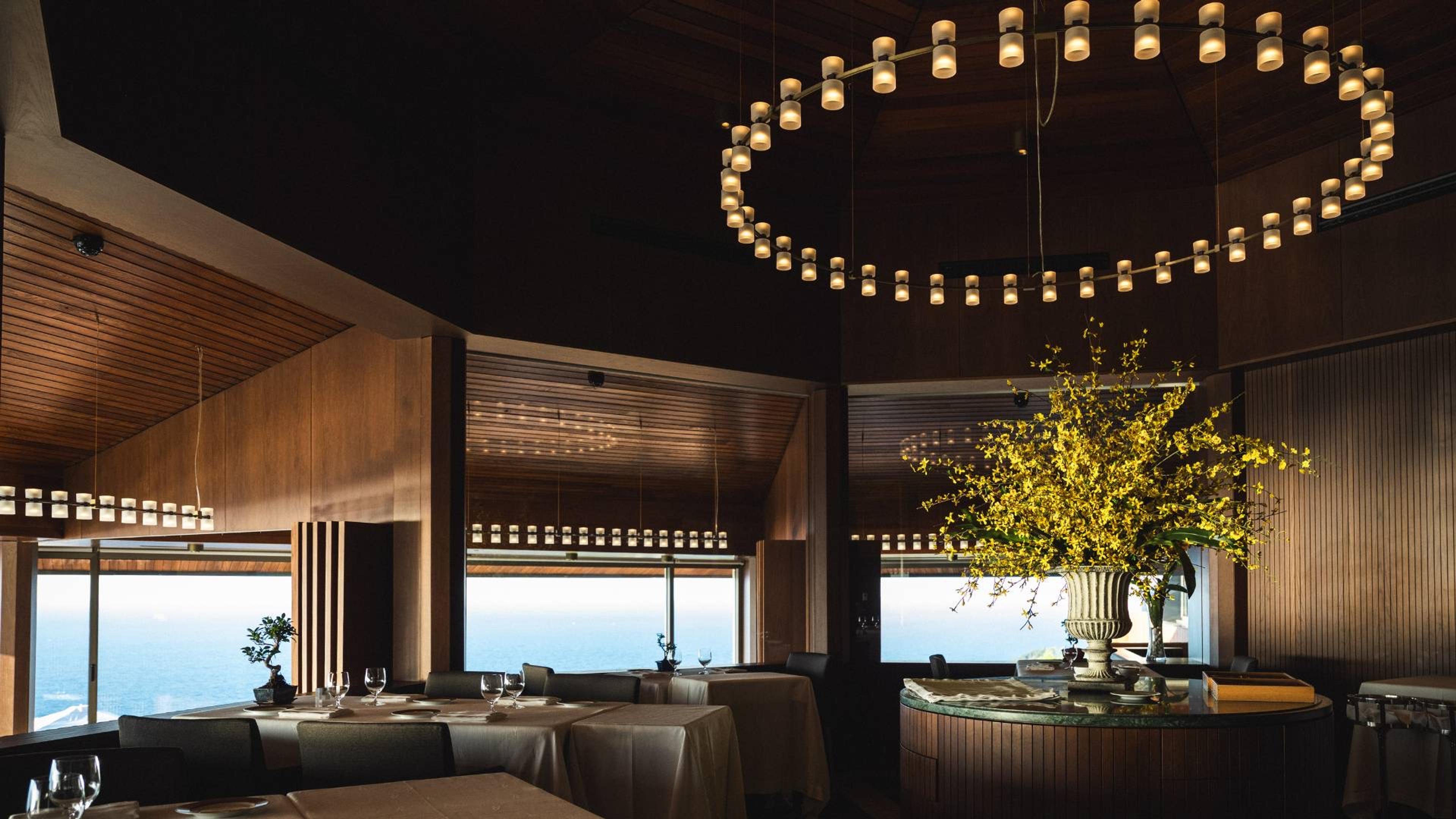 An empty restaurant dining room with white-clothed tables set for dinner, a ring of pendant lights overhead, a tall arrangement of yellow blossom on the bar, and the sea through the windows.