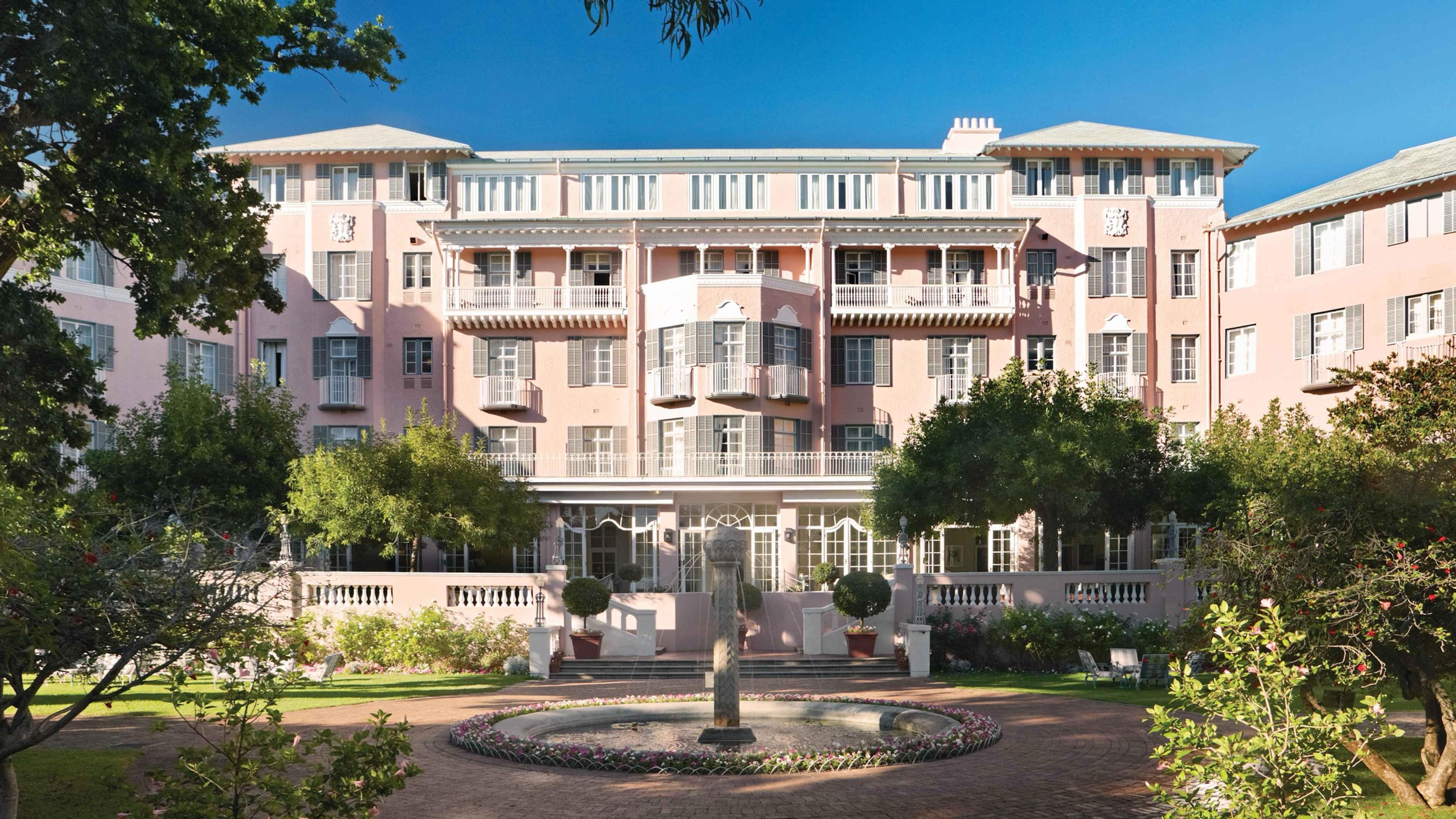 A pink multi-storey hotel with white balconies stands behind a circular fountain and a paved courtyard, with trees on either side under a blue sky.