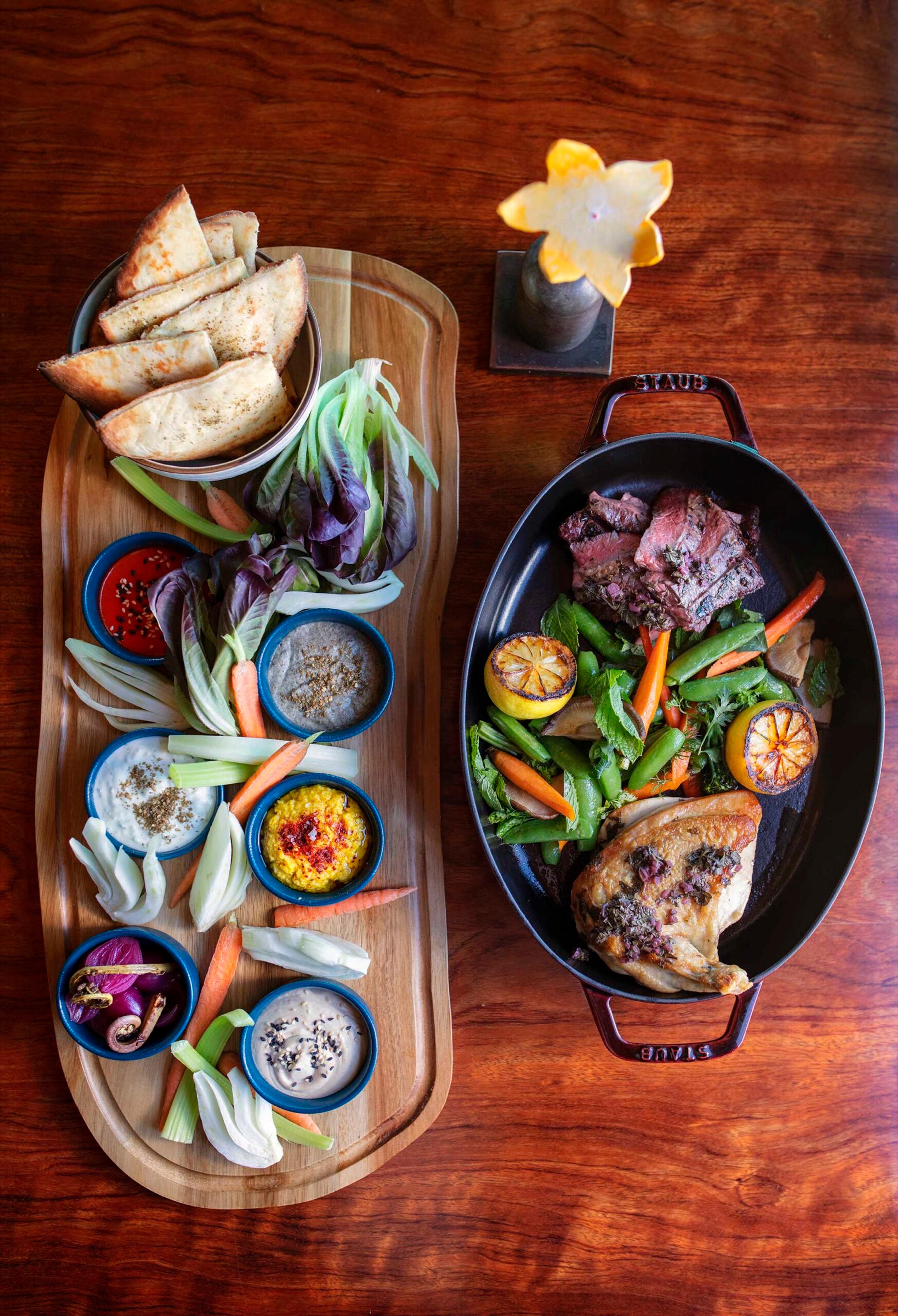 An overhead view of a wooden board with flatbread, raw vegetables and small bowls of dips, beside a cast-iron pan holding sliced steak, roast chicken and vegetables.