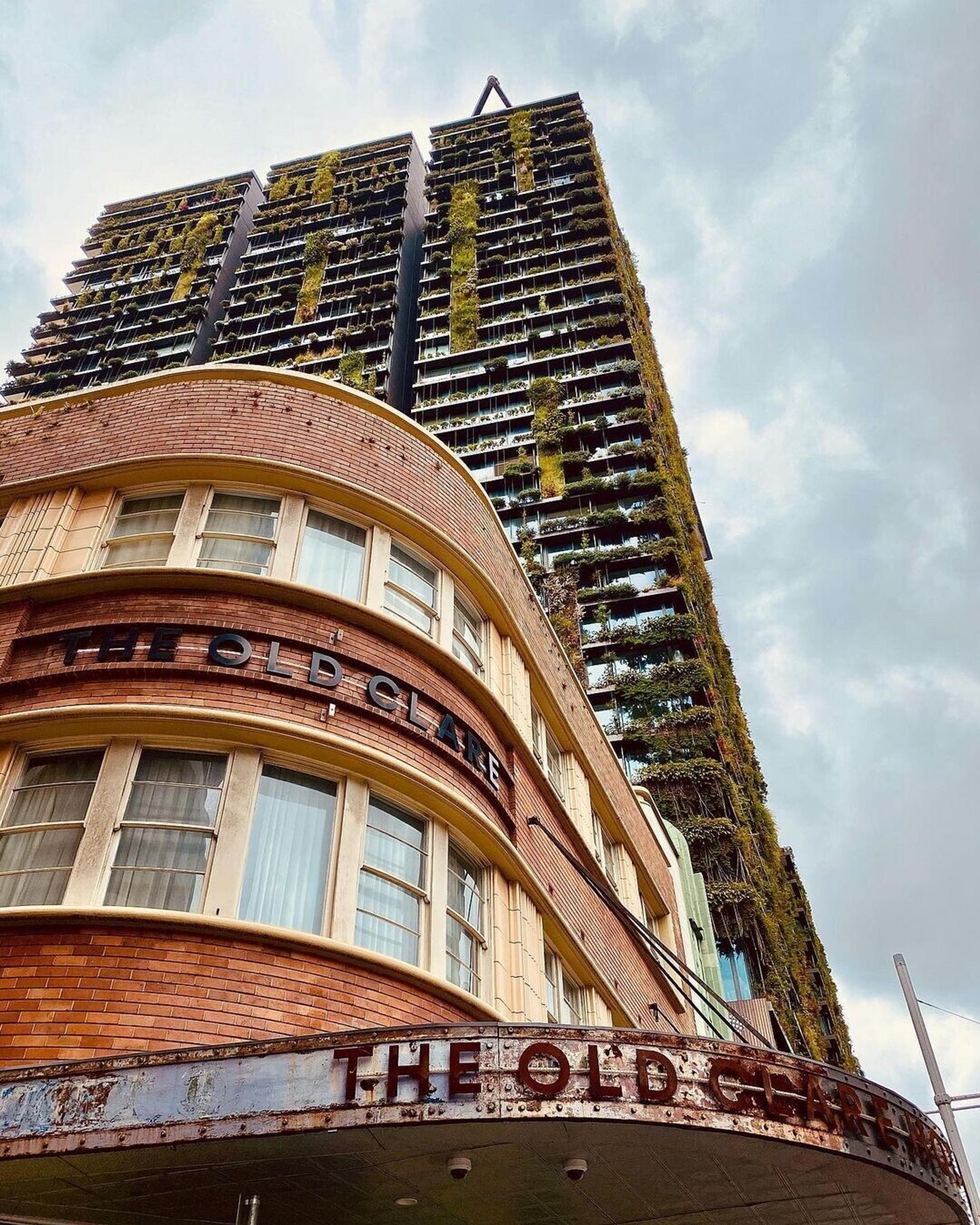 A low angle view of an art deco brick building with the hotel's name on its facade, a glass tower covered in planted greenery rising behind it.