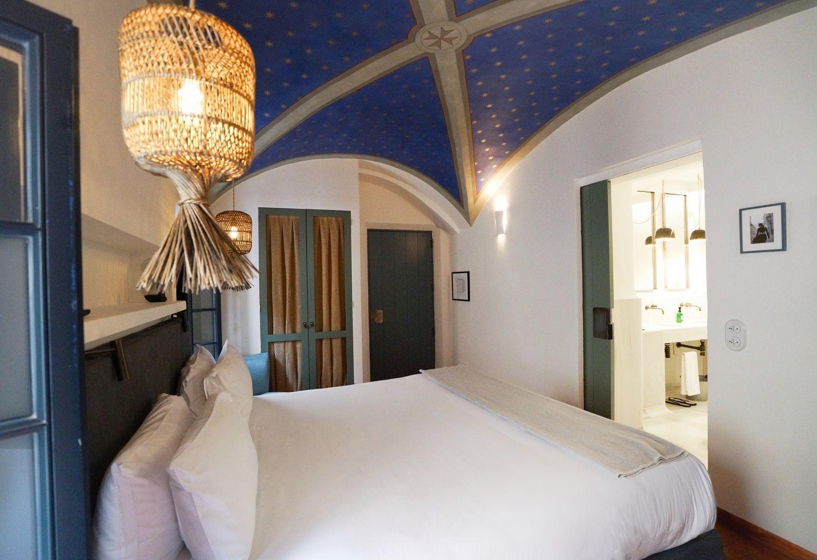 A bed beneath a vaulted ceiling painted dark blue with gold stars, with a bathroom visible through an open doorway.