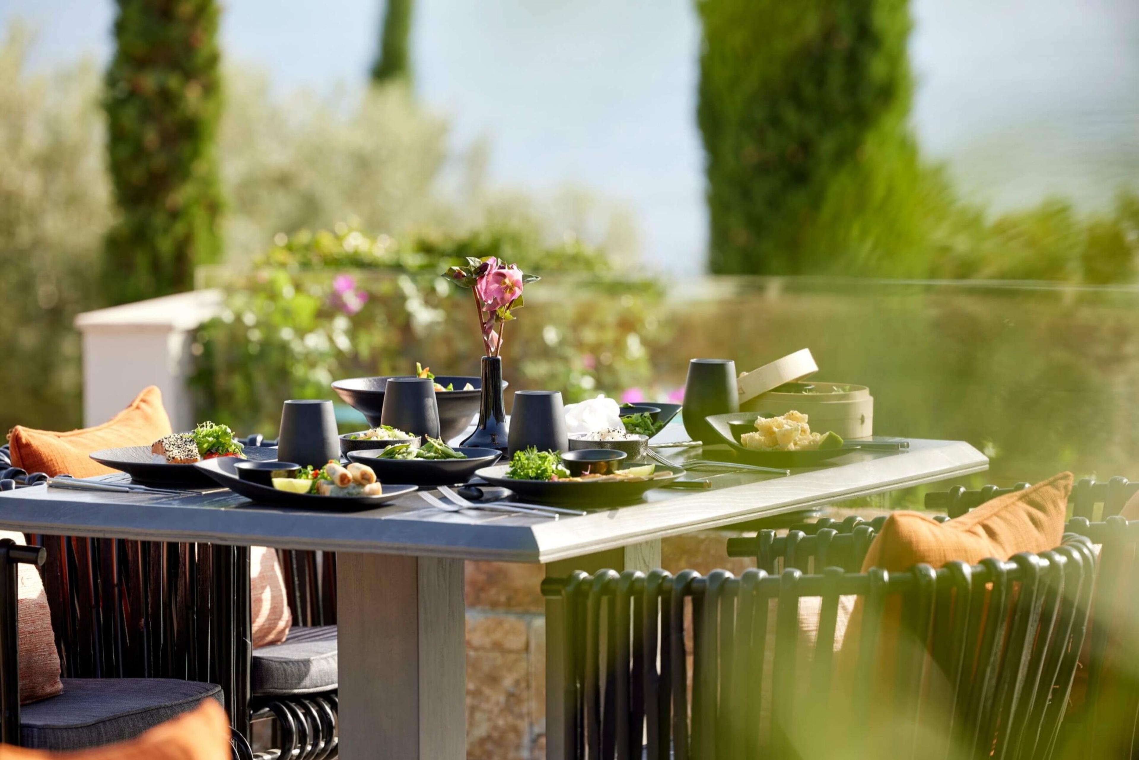 An outdoor dining table set with small plates of food and a vase of flowers, shaded by pine trees with the sea visible beyond.