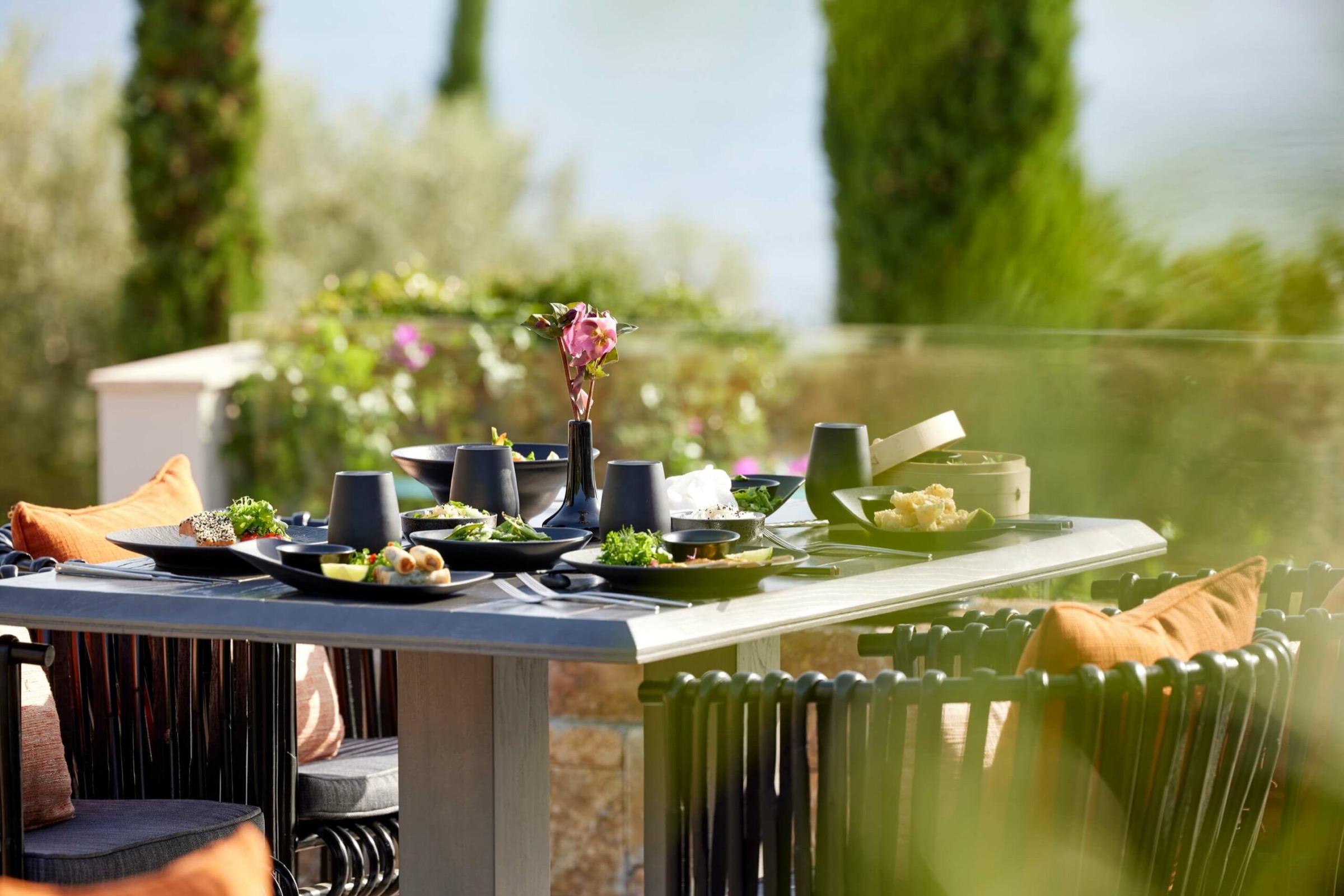 An outdoor dining table set with small plates of food and a vase of flowers, shaded by pine trees with the sea visible beyond.