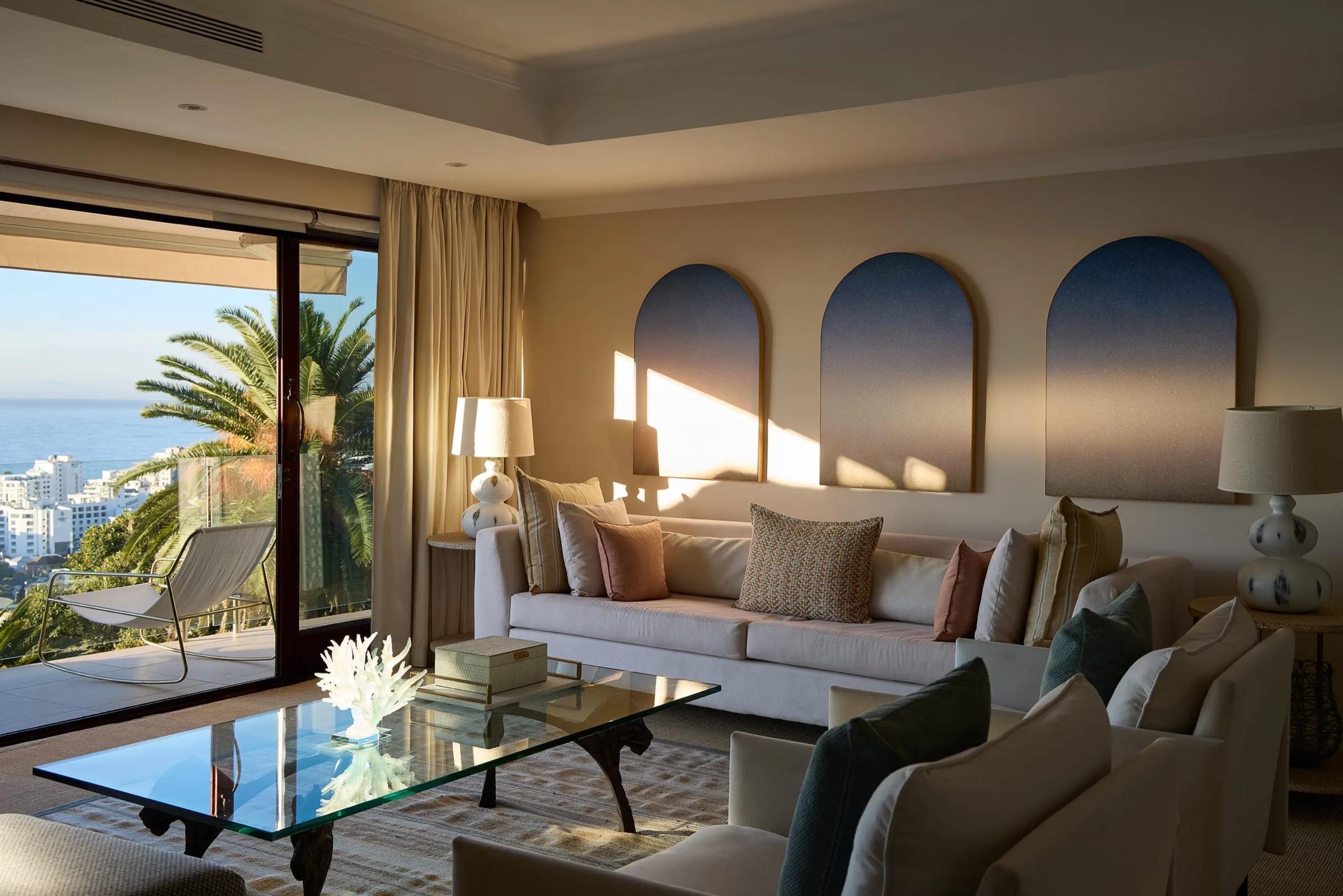A suite living room with three arched blue canvases on the wall, a pale sofa and a glass coffee table, with sliding doors open onto a terrace, a palm and the sea beyond.