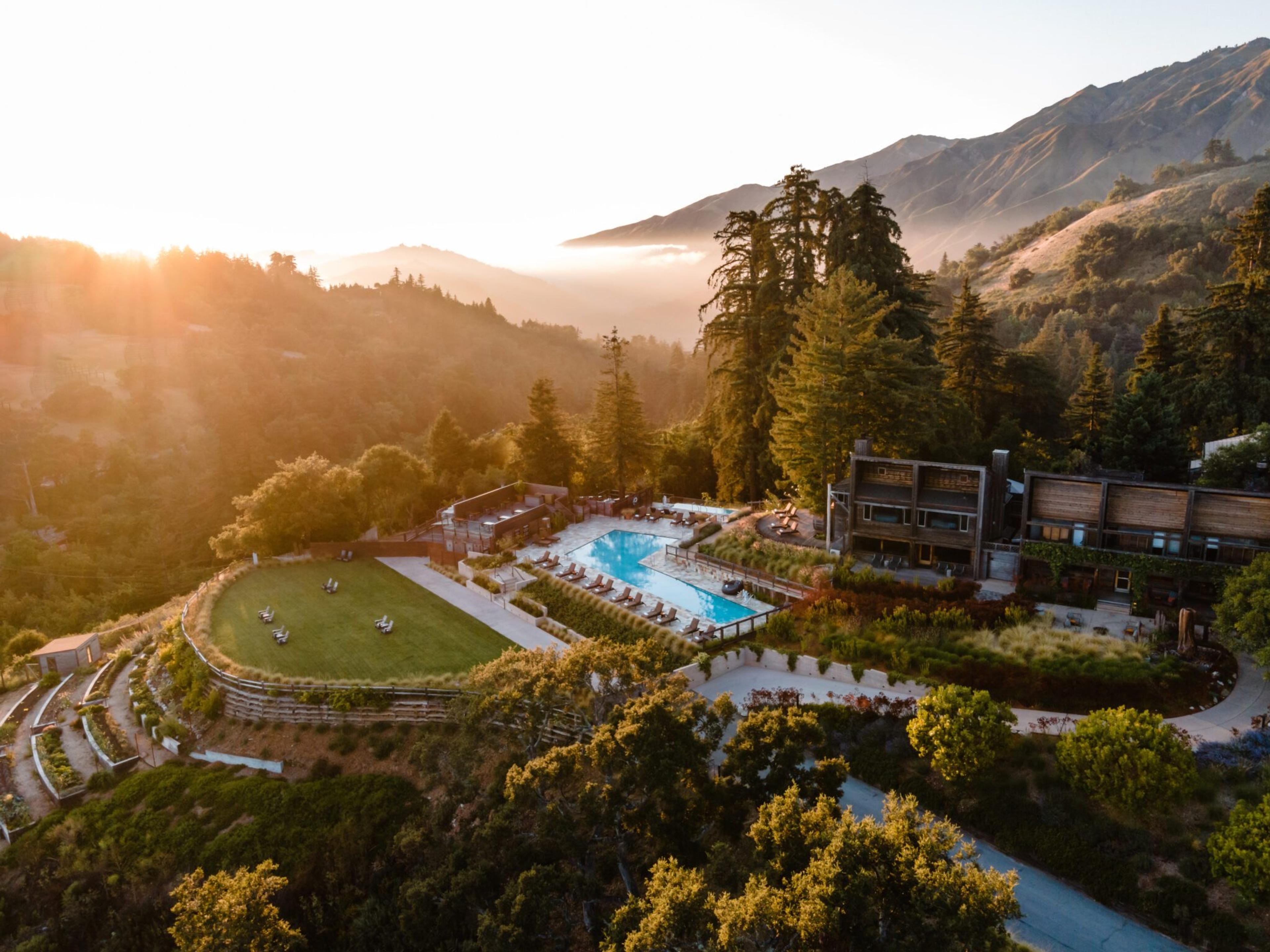 an aerial view at sunrise of a hillside resort with two swimming pools, a lawn and wood-clad buildings surrounded by redwood forest and mountains