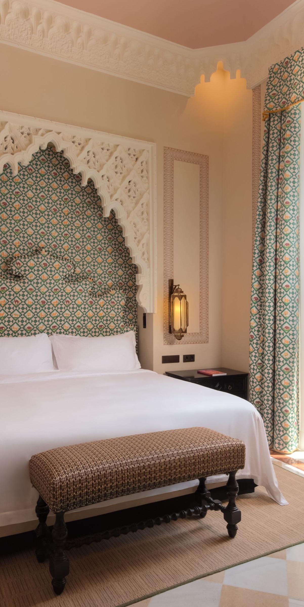 A bedroom with a carved plaster arch framing a headboard upholstered in a green and cream pattern of animals, a lit brass lantern beside it and a woven bench at the foot of the bed.