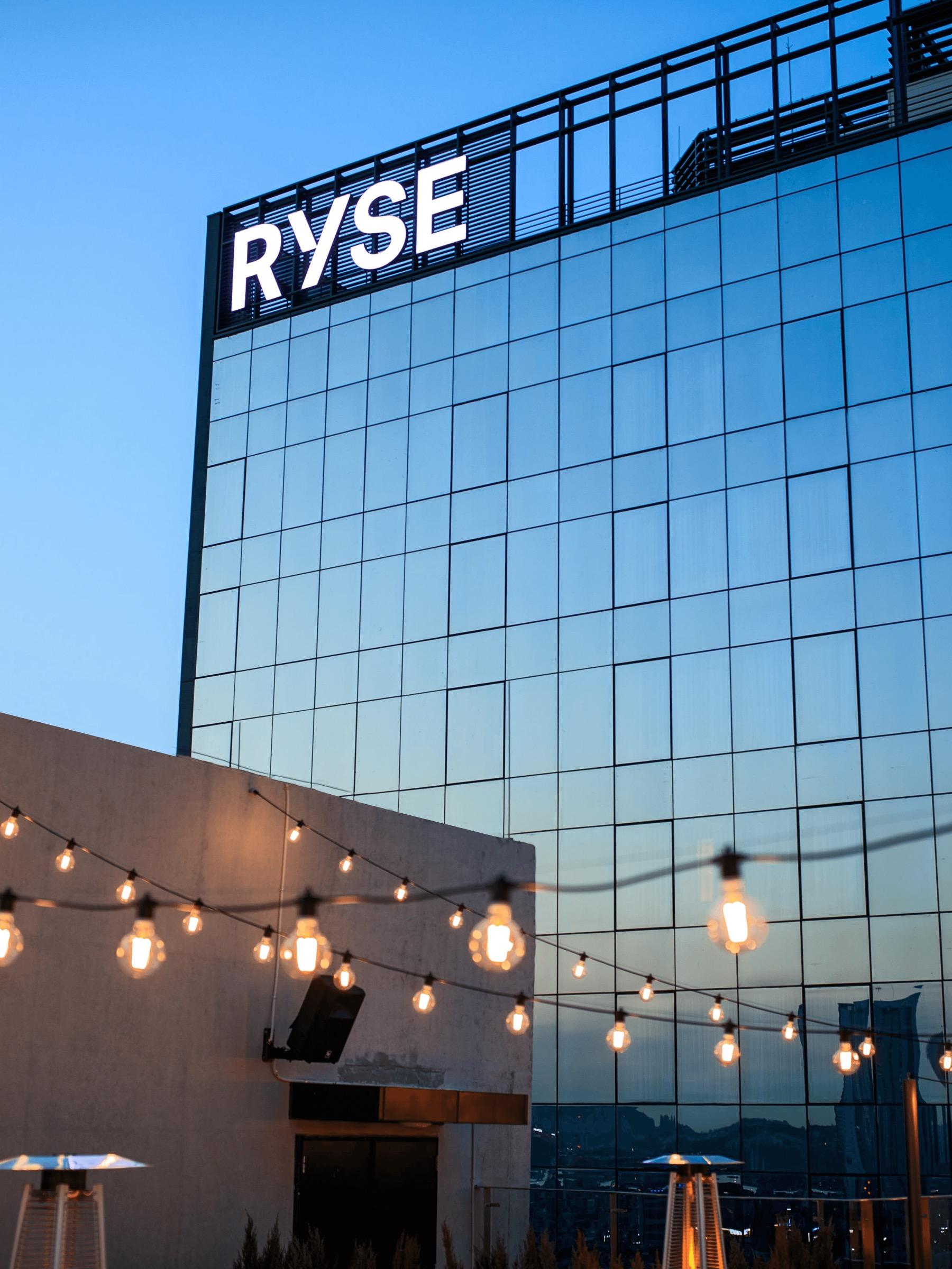 The glass tower of RYSE Hotel lit at dusk with its sign on the roofline, string lights strung above a rooftop terrace in the foreground.