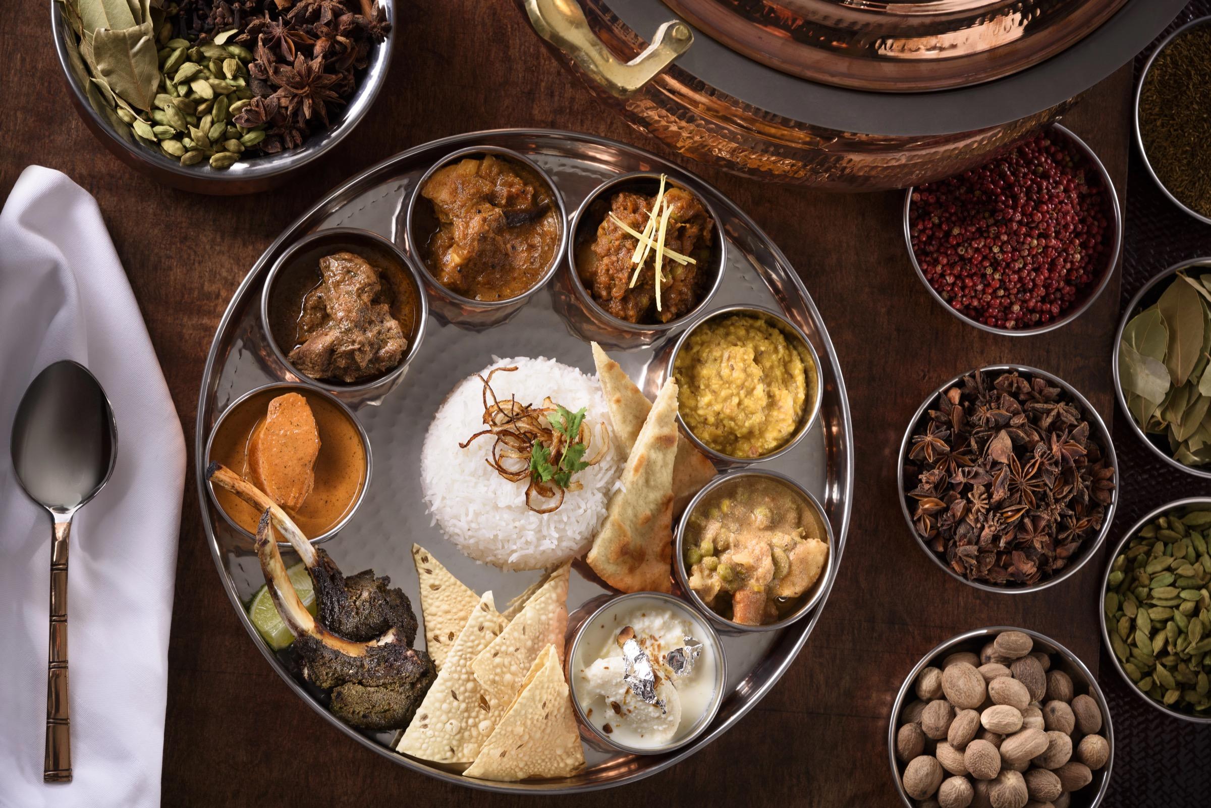 An overhead shot of a shared Indian-style feast on steel dishes: curries, rice, lamb chops and naan, with a spoon and poppadoms beside it.