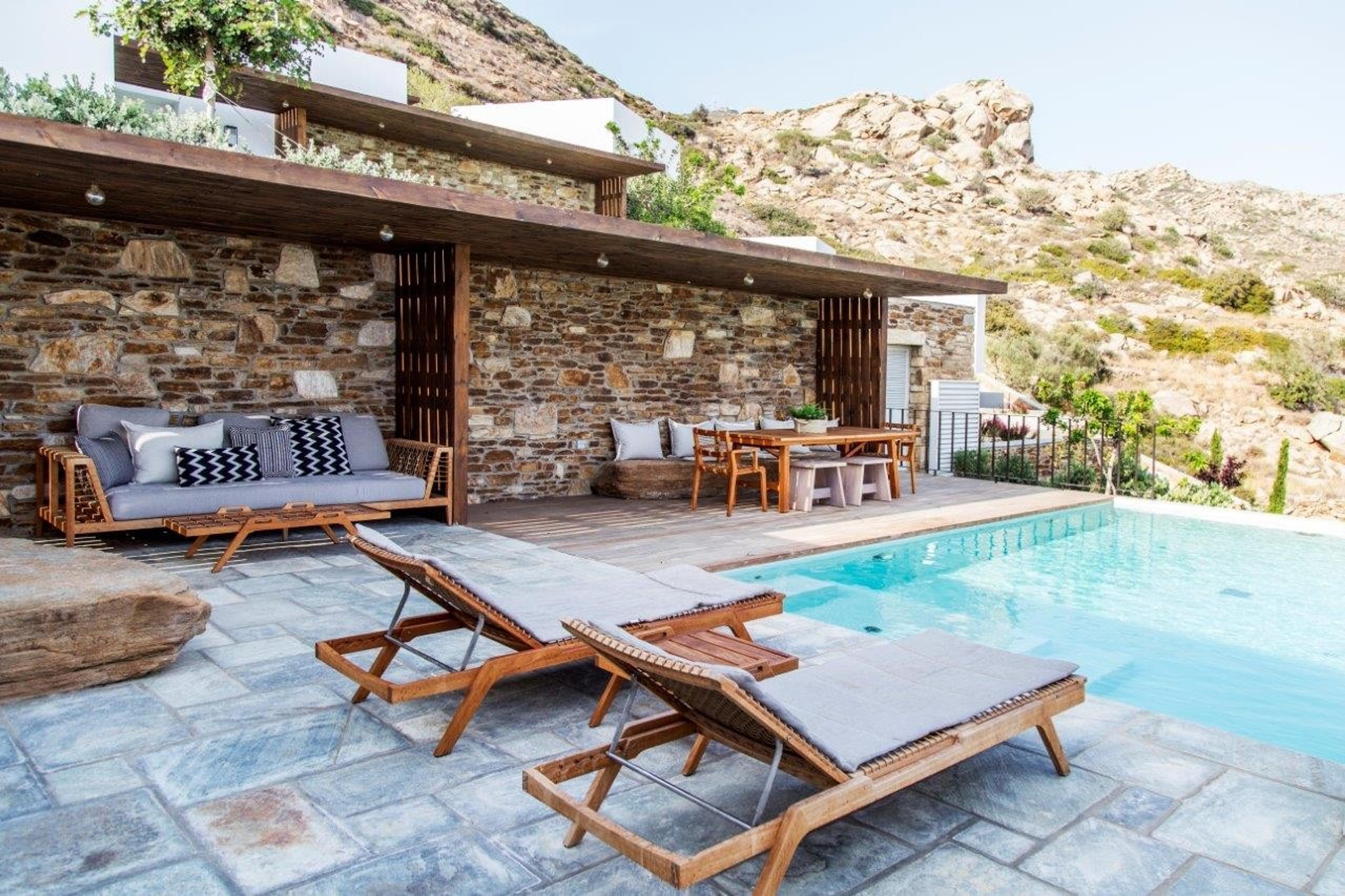 A pool beside a timber-roofed stone terrace, with sofas, loungers and a dining table under the pergola, and rocky hills behind.