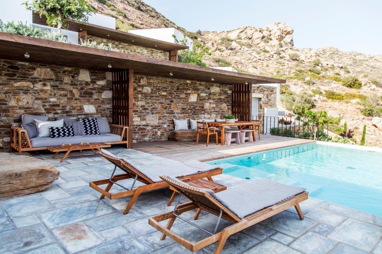 A pool beside a timber-roofed stone terrace, with sofas, loungers and a dining table under the pergola, and rocky hills behind.