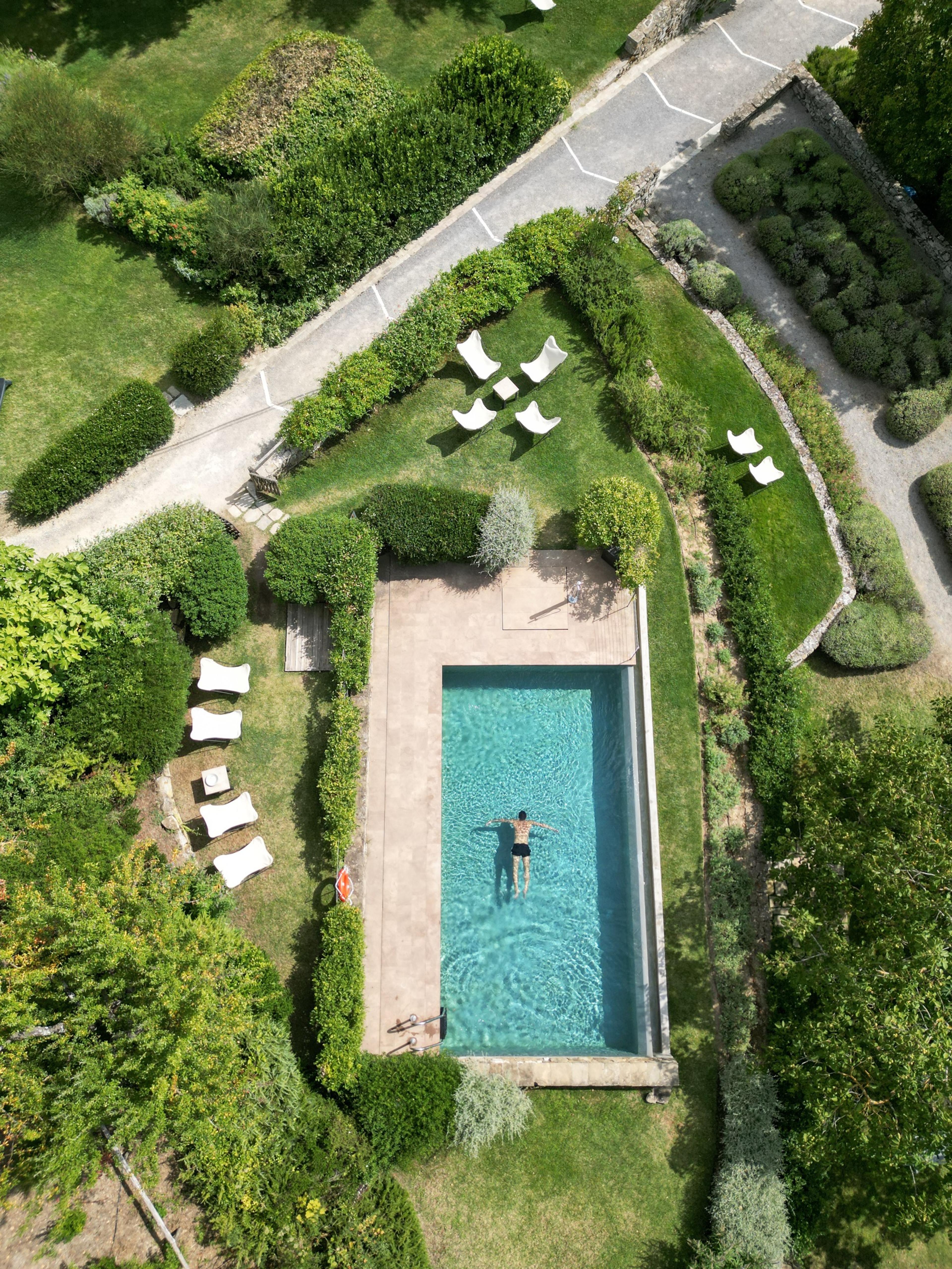 An aerial view of a rectangular swimming pool in a garden, a person floating on their back in the water with sun loungers set around the edge.