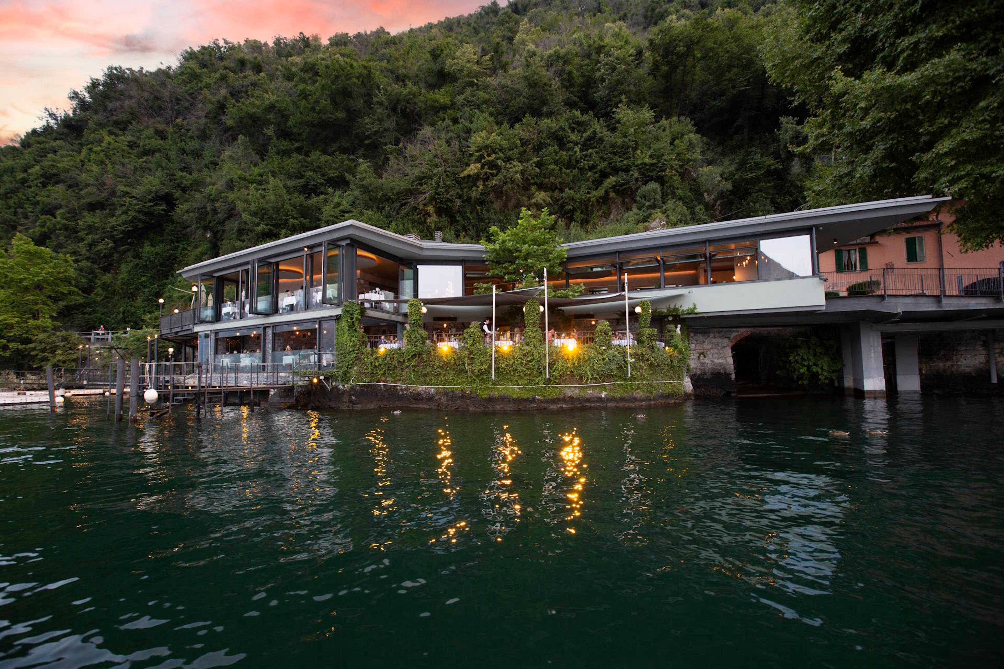 A lakeside restaurant with a covered terrace over the water, wooded hills rising behind it