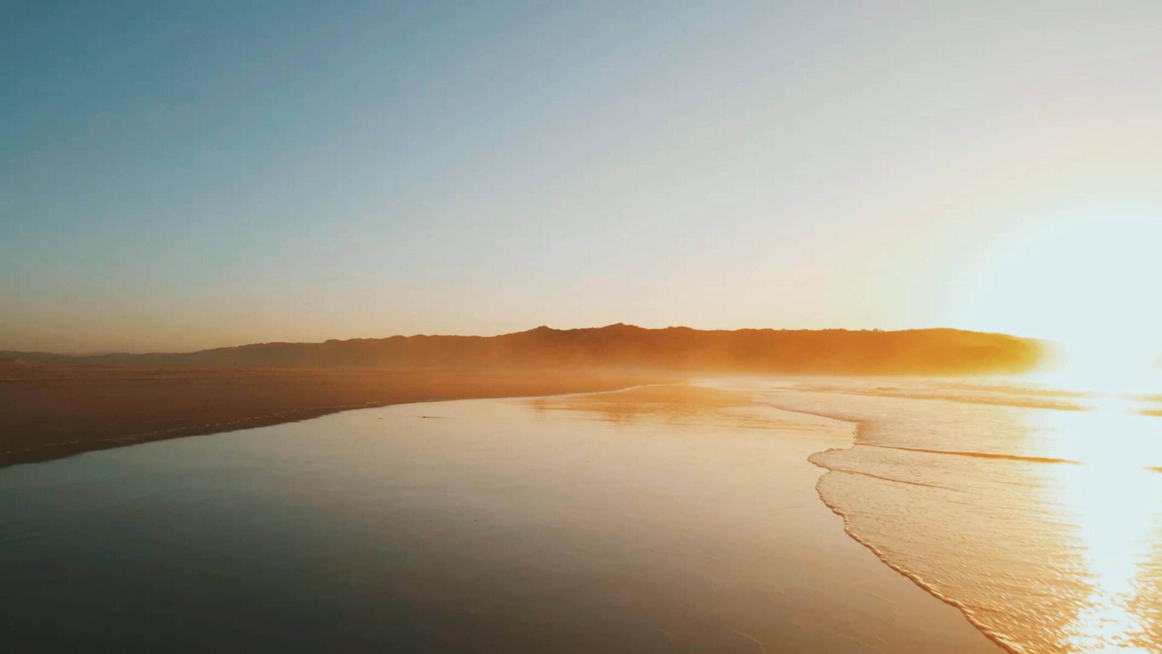 A wide, calm beach at sunrise with low hills in the distance and the sun reflecting on wet sand.