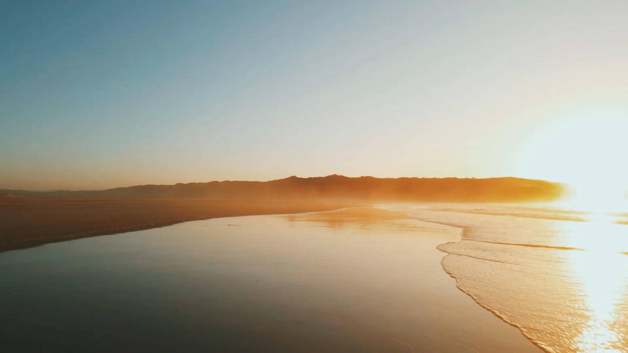 A wide, calm beach at sunrise with low hills in the distance and the sun reflecting on wet sand.