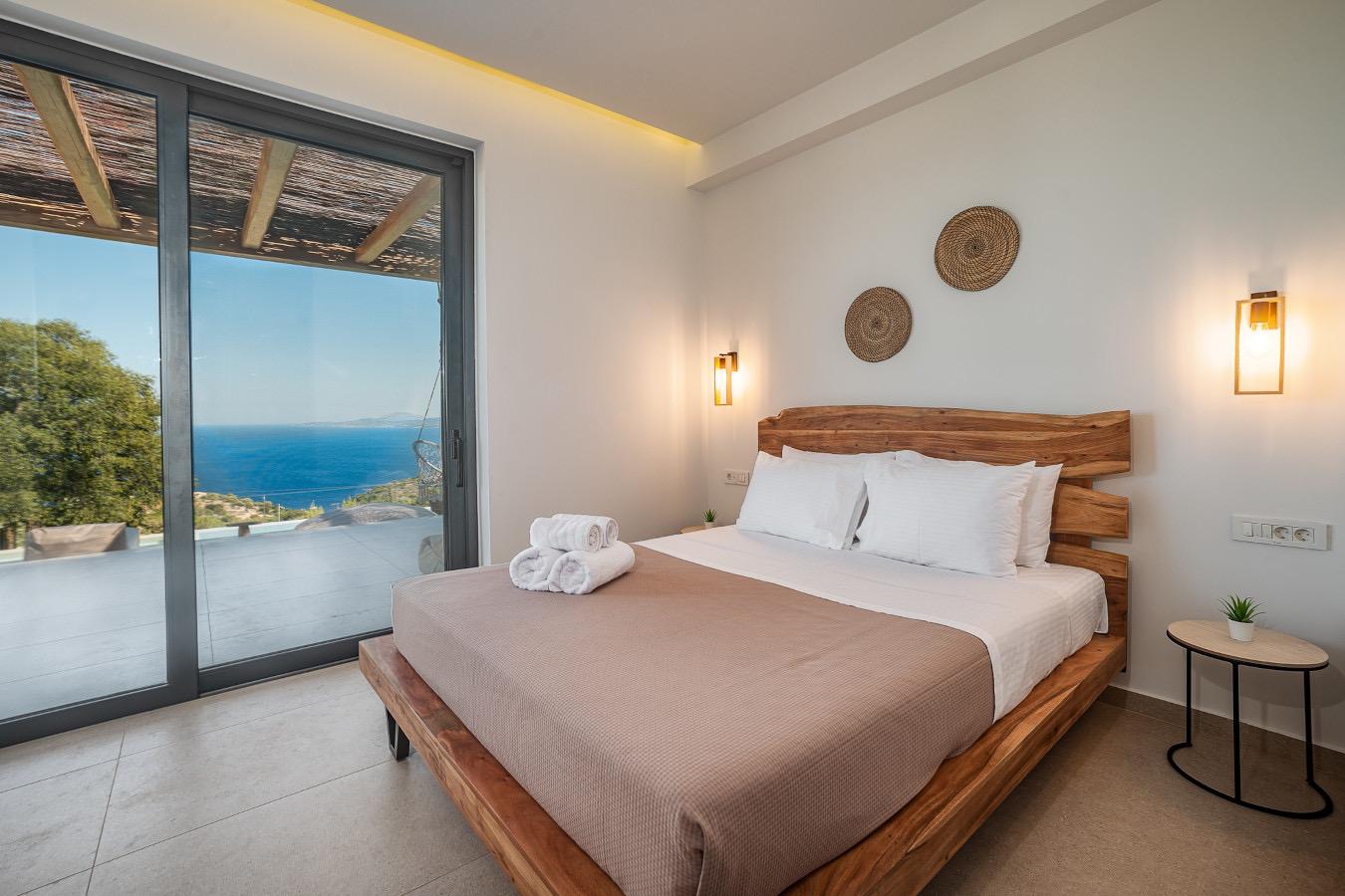 A bedroom with a pale wooden bed frame, white pillows and rolled towels, with a sliding glass door open to a sea view and a pergola outside.
