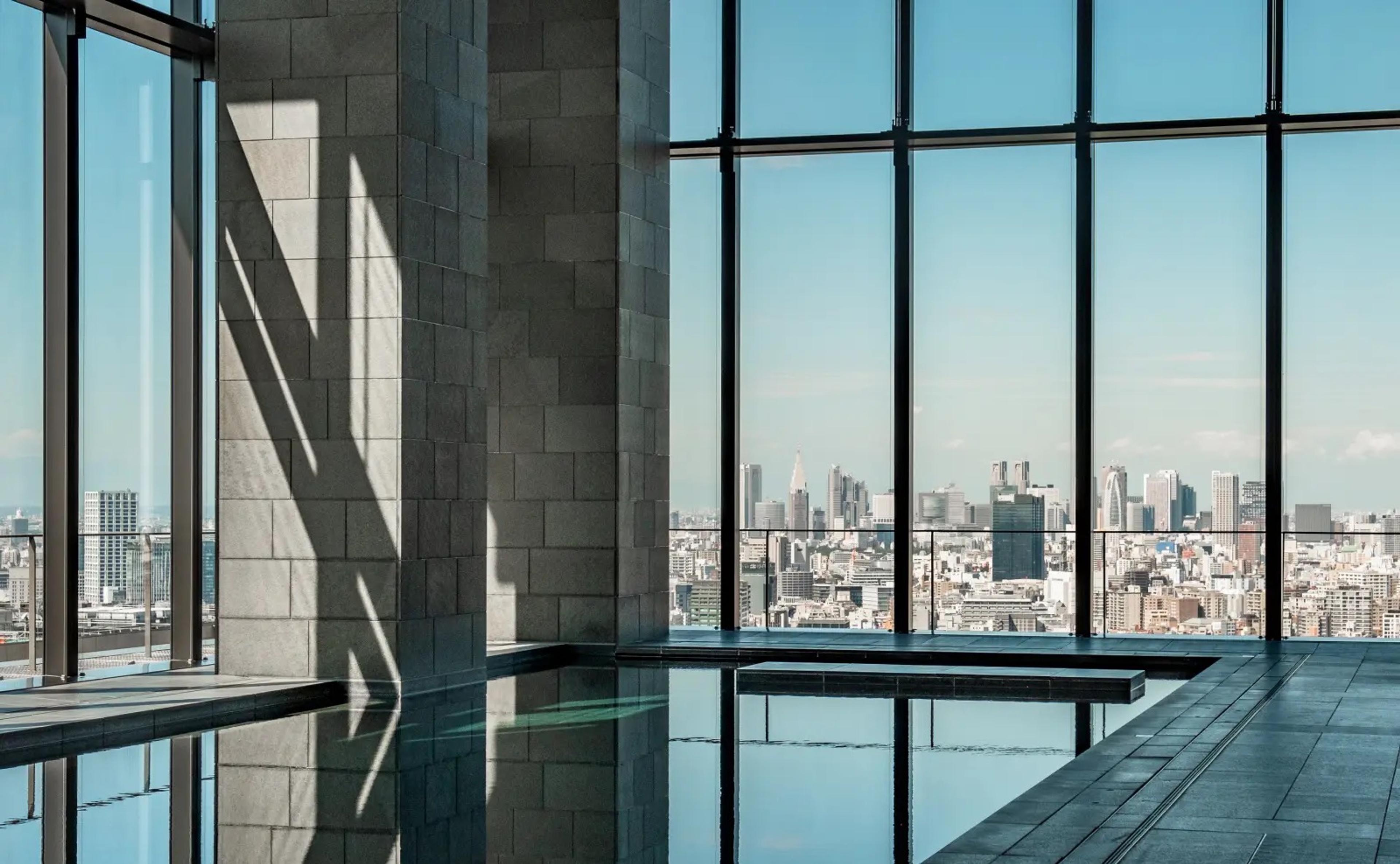 An indoor pool lit by daylight, running alongside floor-to-ceiling windows with the Tokyo skyline beyond.