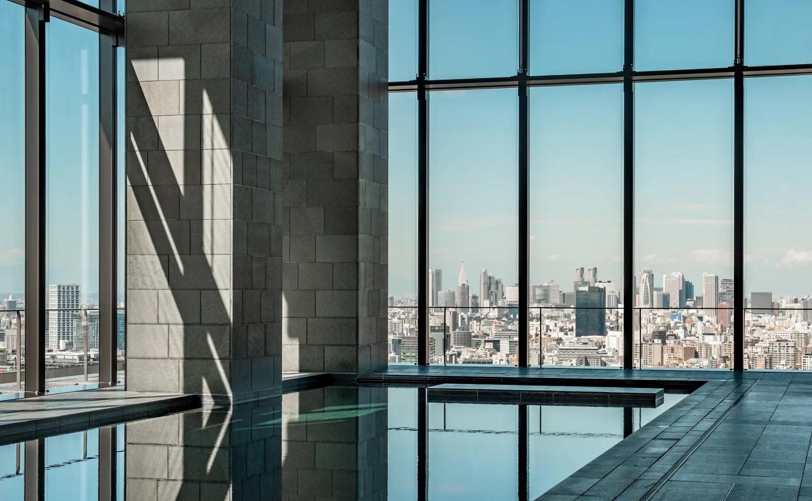An indoor pool lit by daylight, running alongside floor-to-ceiling windows with the Tokyo skyline beyond.
