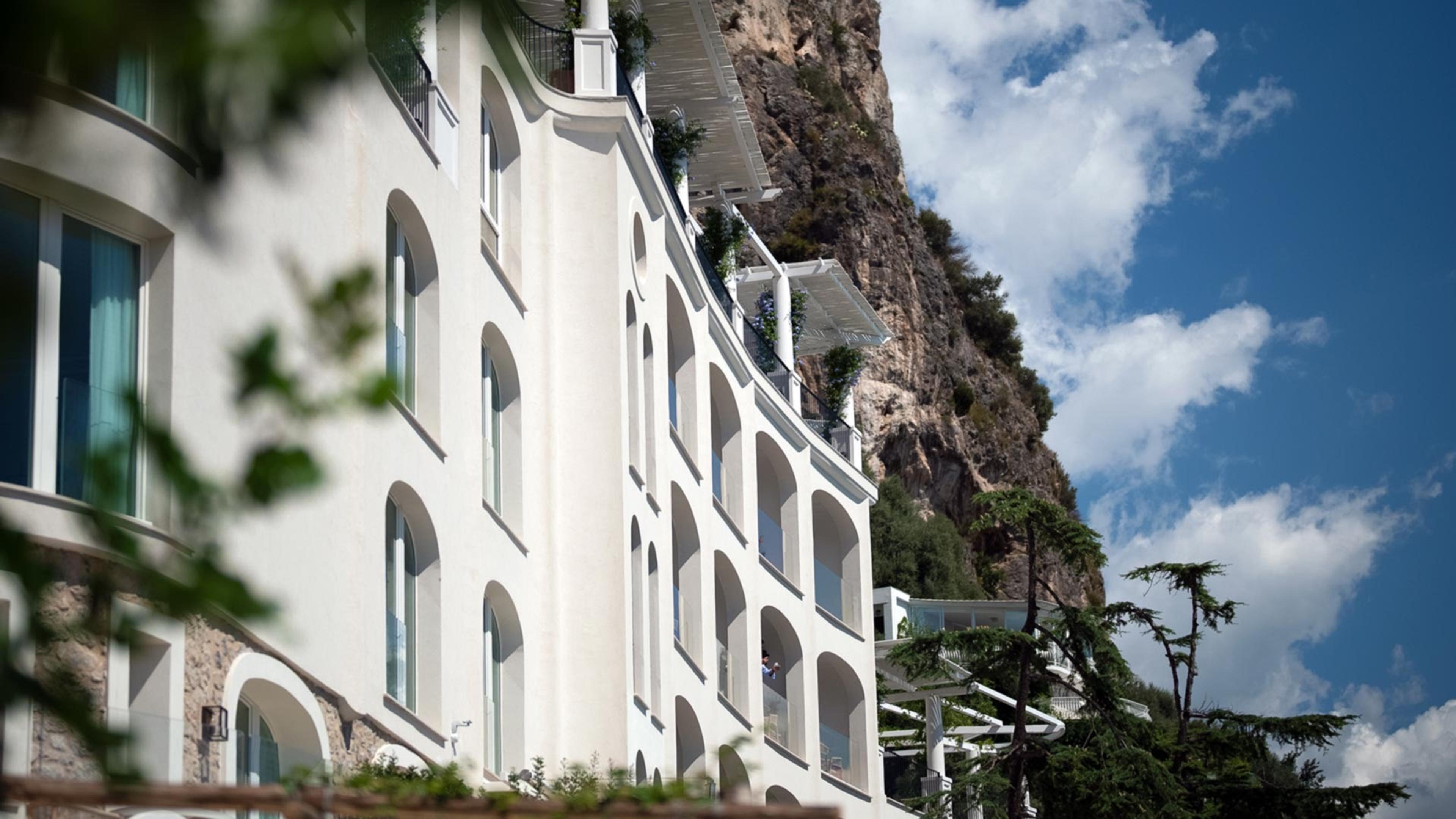 A curved white hotel building with arched balconies set against a sheer rock cliff, photographed from below through out-of-focus leaves.