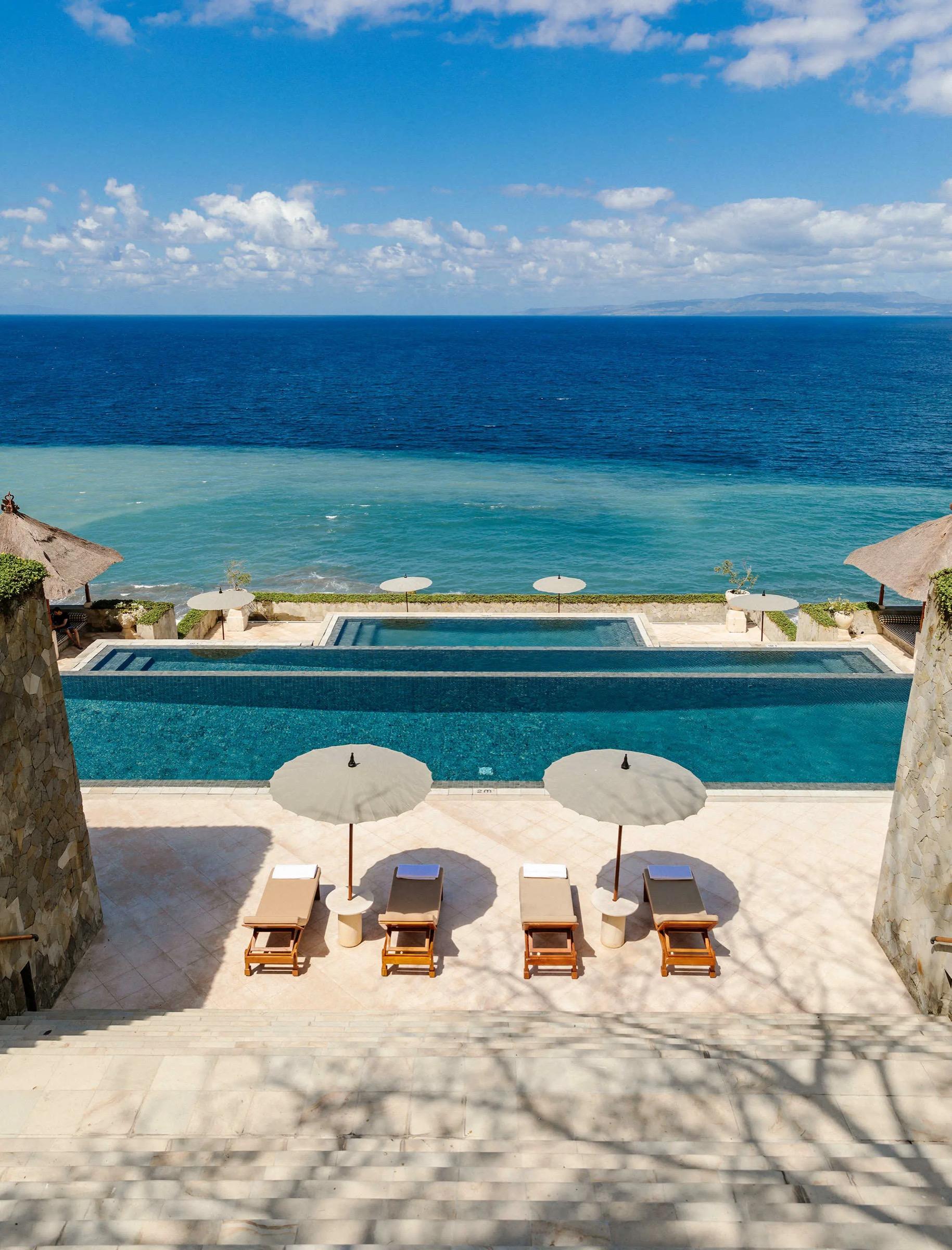 A set of tiered pools steps down a terrace toward the sea, with sun loungers and parasols along the edge under a blue sky.