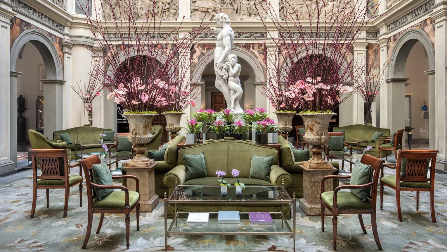 A courtyard seating area with green sofas and armchairs, tall floral arrangements and a classical statue of two figures under an arcade.