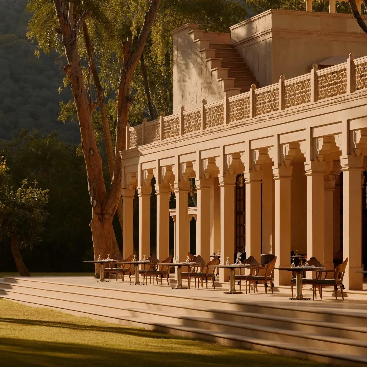 An outdoor dining terrace with set tables and chairs along a colonnade, lit by low evening sun with trees and steps beyond.