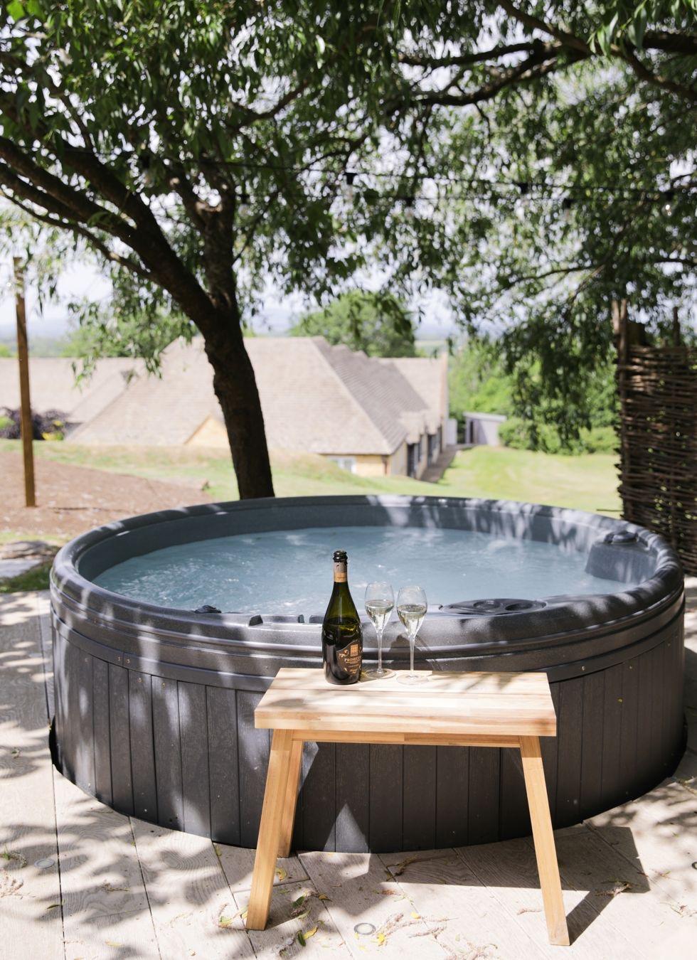 A round outdoor hot tub shaded by a tree, with a bottle of champagne and two glasses set on a small wooden stool, cottages visible through the branches beyond.