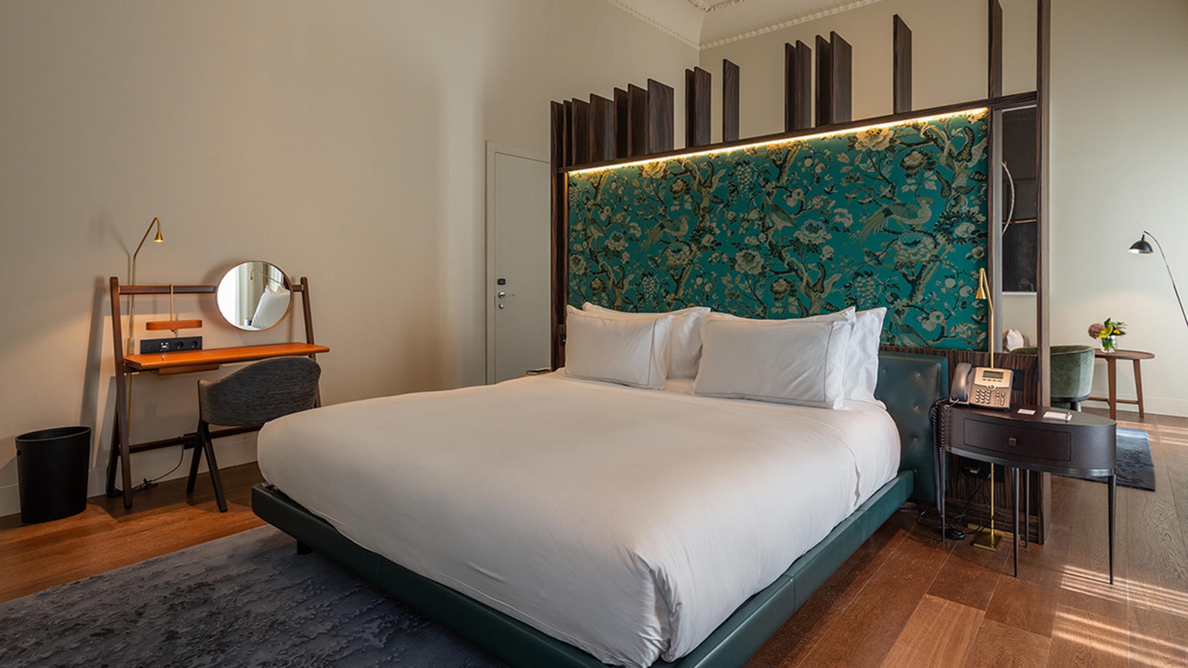 A bed with a teal floral fabric panel behind the headboard sits between a mirrored dressing table on one side and a dark side table holding a telephone on the other, on a grey rug over wood flooring.