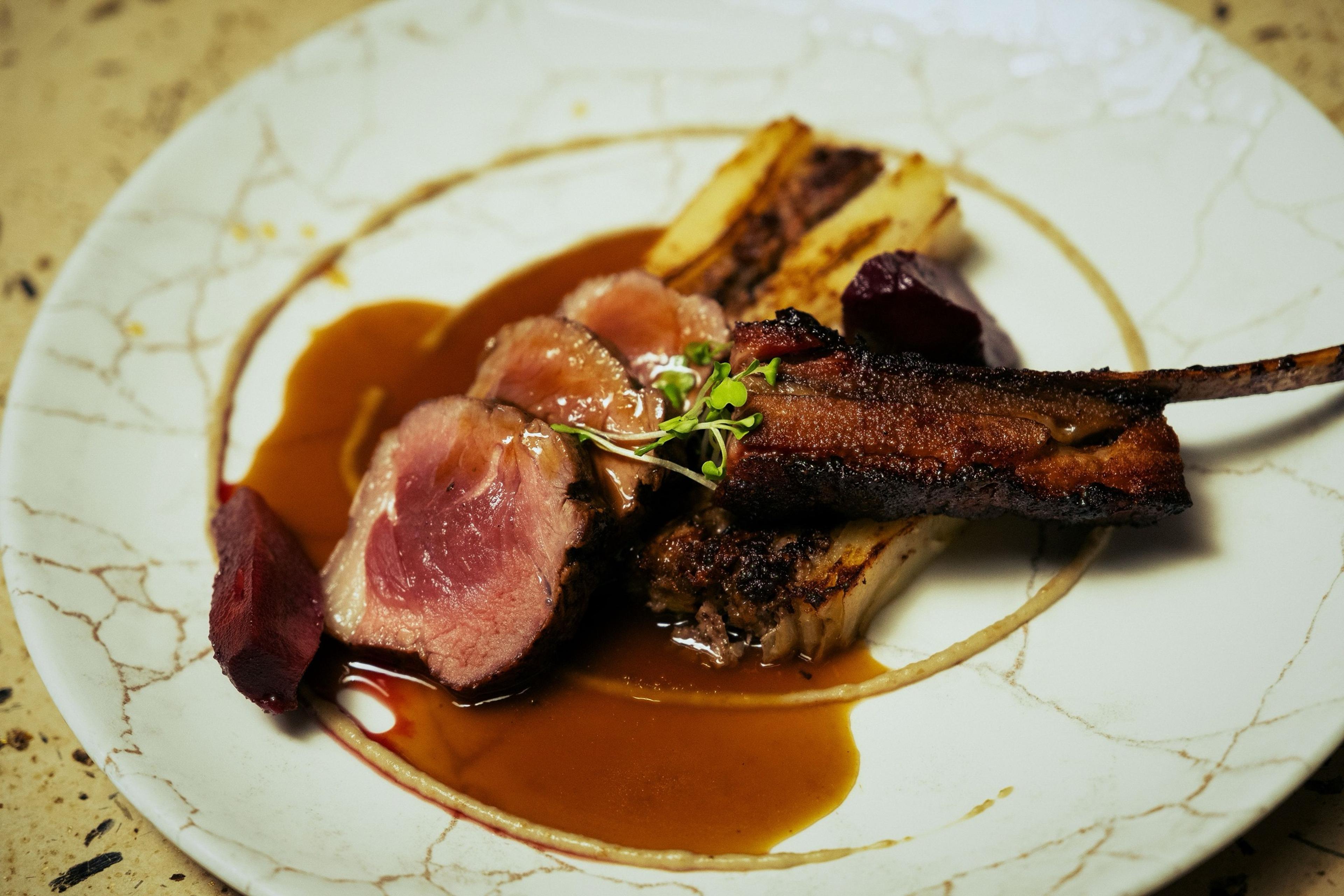 A white plate with sliced pink roast meat, a charred rib, roasted vegetables and a pool of dark sauce.