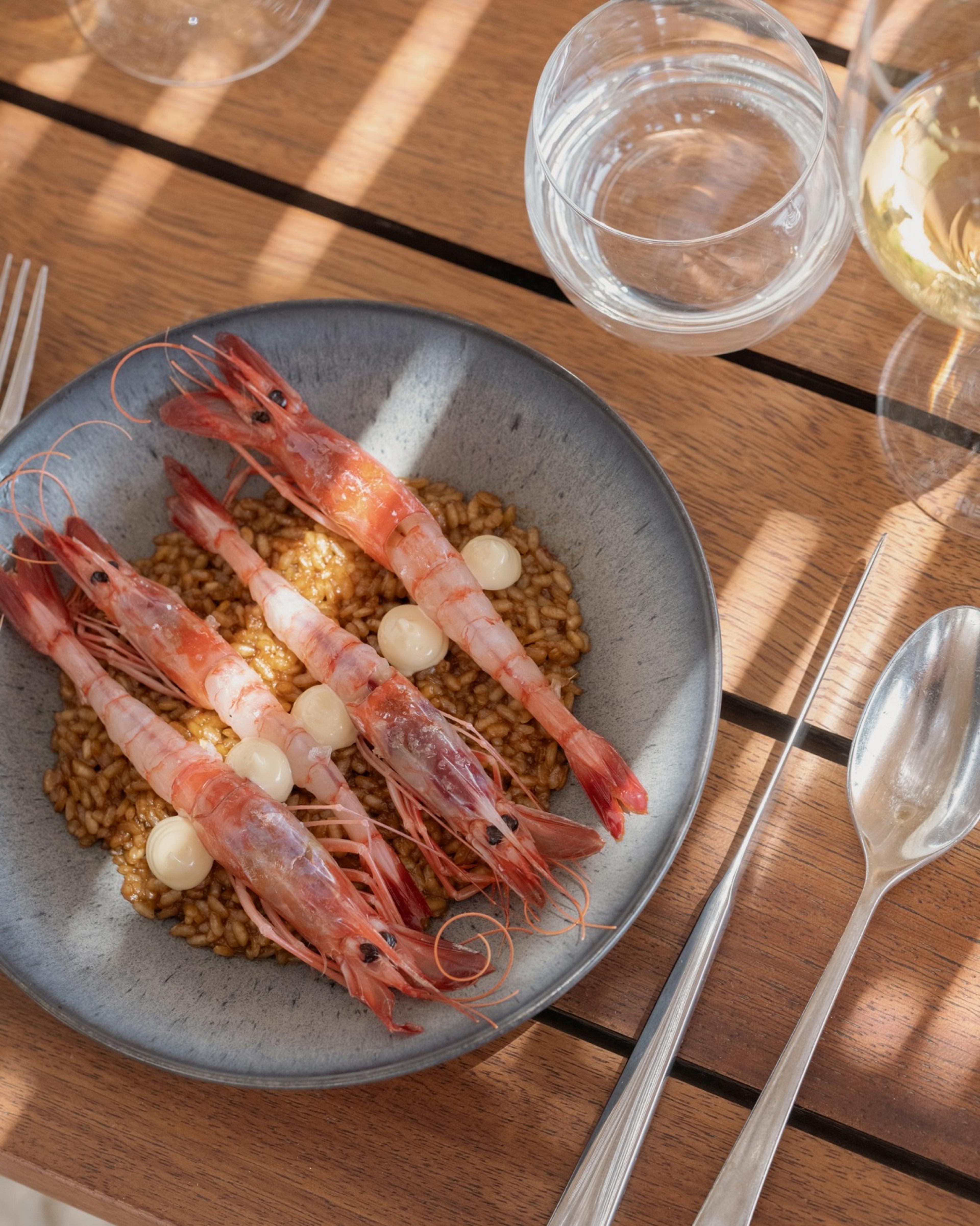 A plate of whole prawns on rice, with wine and water glasses, a fork and two spoons on a wooden outdoor table.