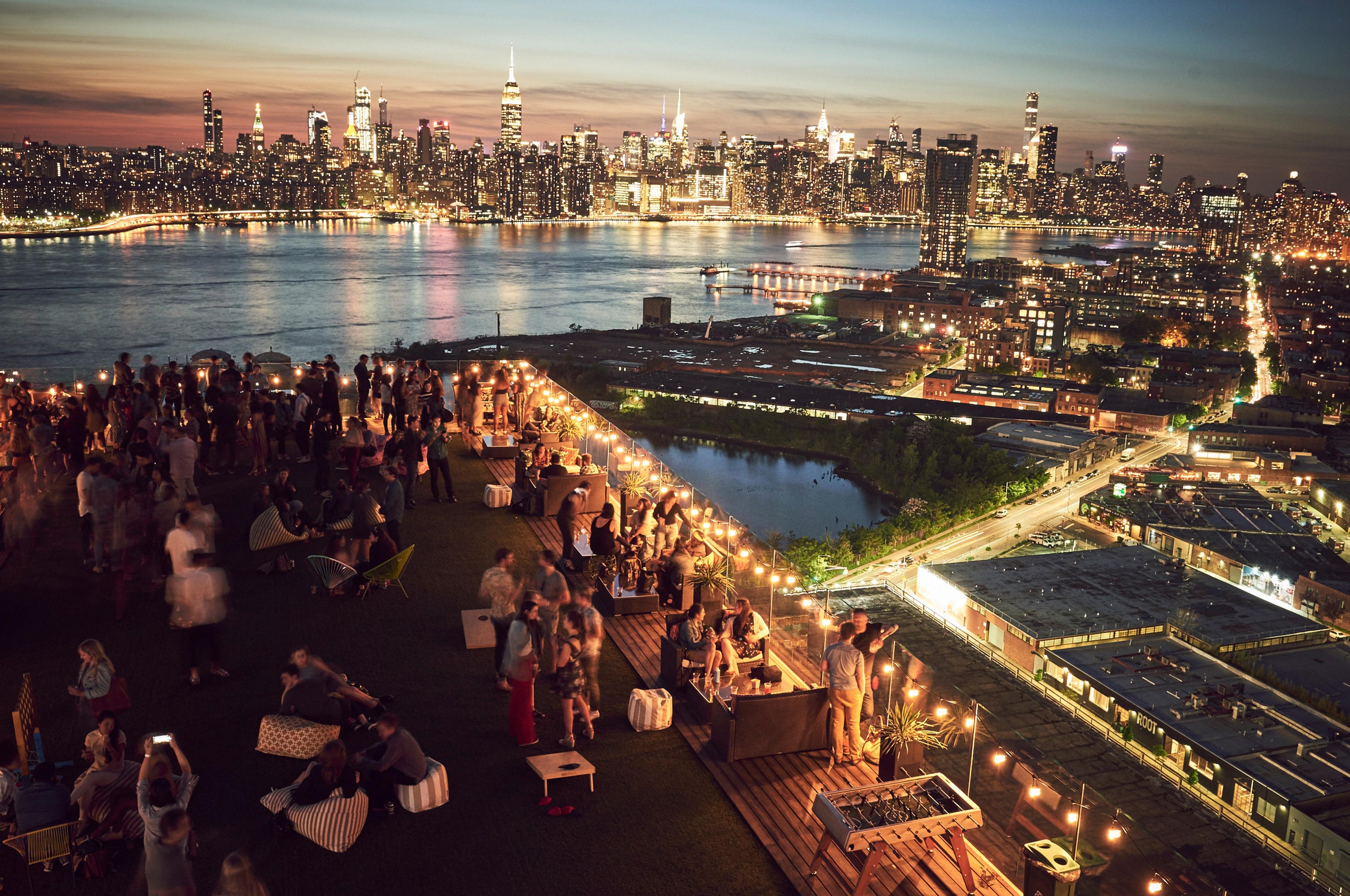 An aerial view of a rooftop party at night, with the crowd lit by string lights and the Manhattan skyline glowing across the river.