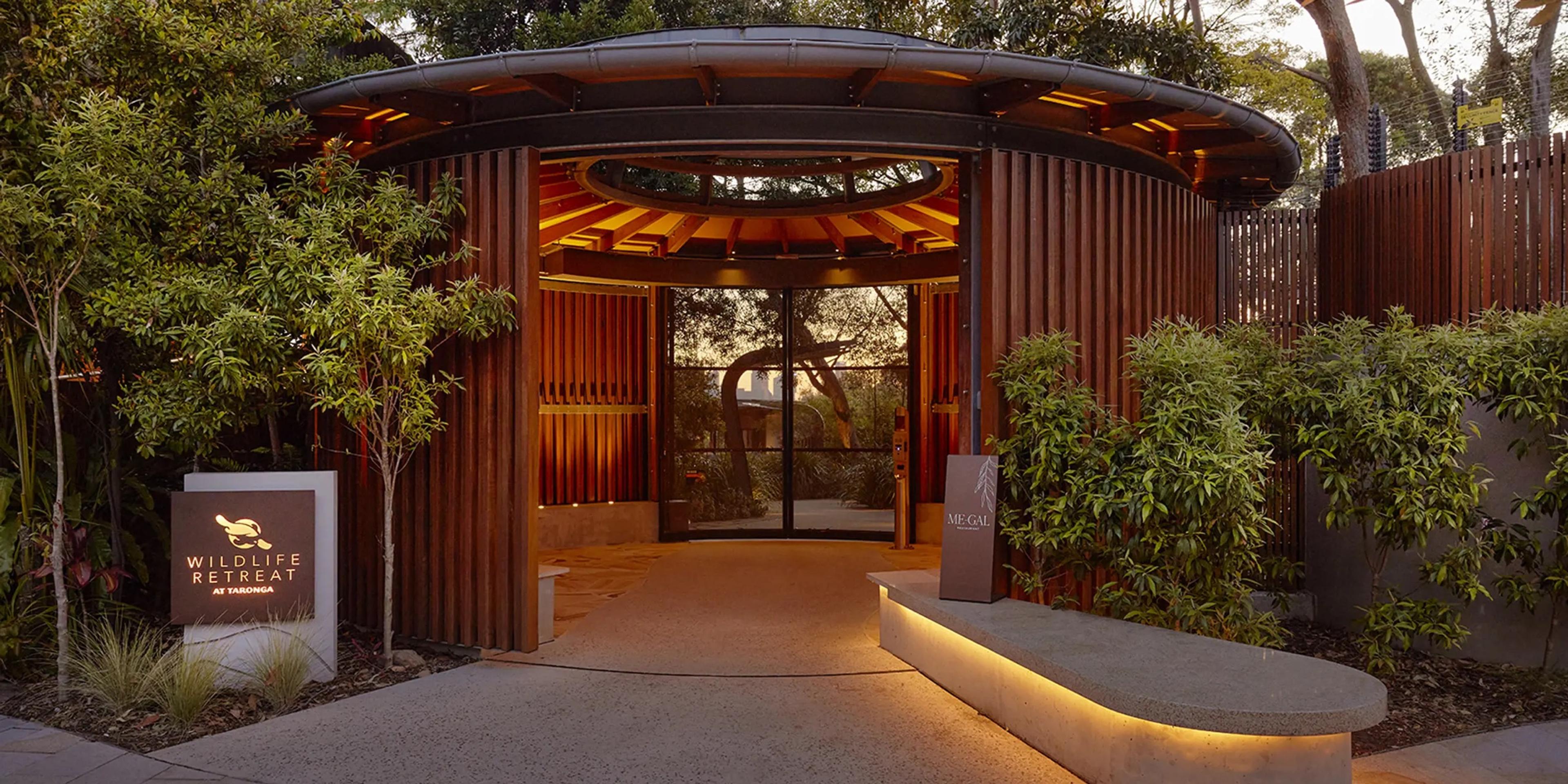A timber-slatted entrance canopy lit at dusk, with a sign reading Wildlife Retreat at Taronga beside the path and glass doors open at the far end.