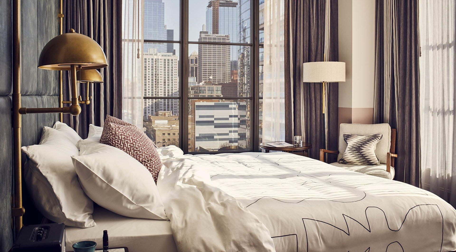 A hotel bed with a charcoal panelled headboard and brass lamp beside a window looking over the Chicago skyline at dusk.