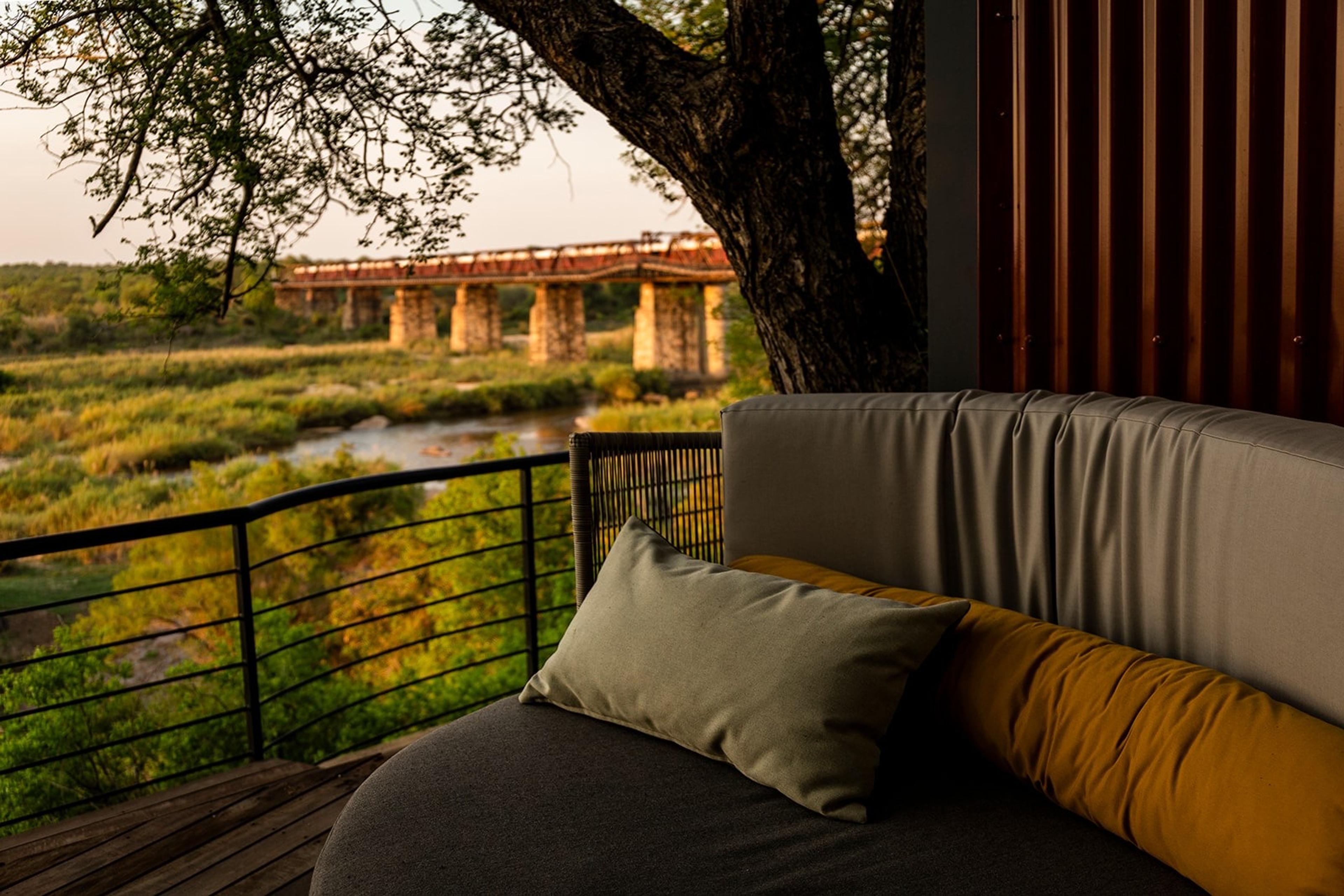 An outdoor daybed with cushions under a tree on the hotel's deck, with the steel bridge and a train crossing it visible in the distance.