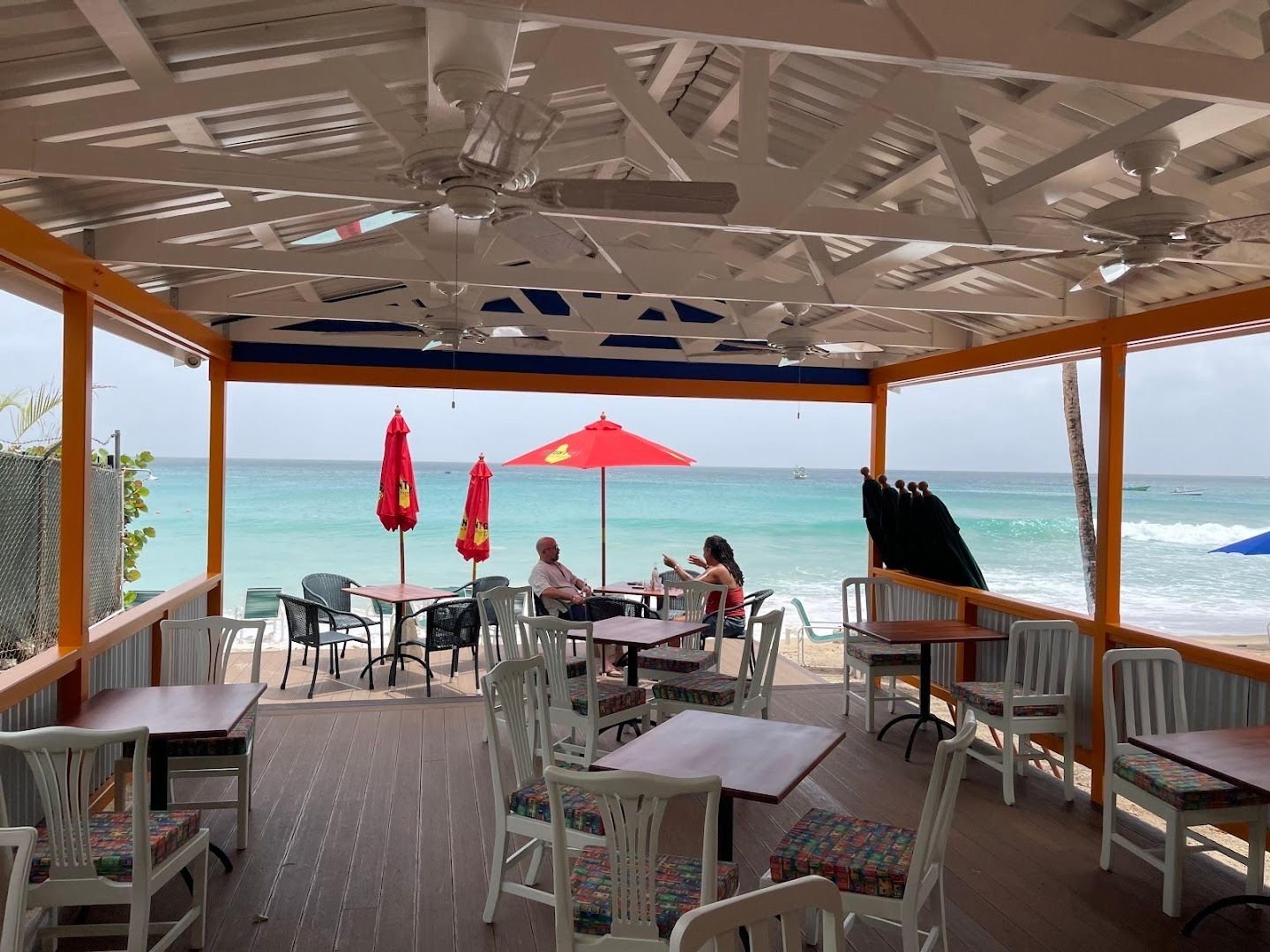 A covered beach terrace of tables under red umbrellas, the sea beyond