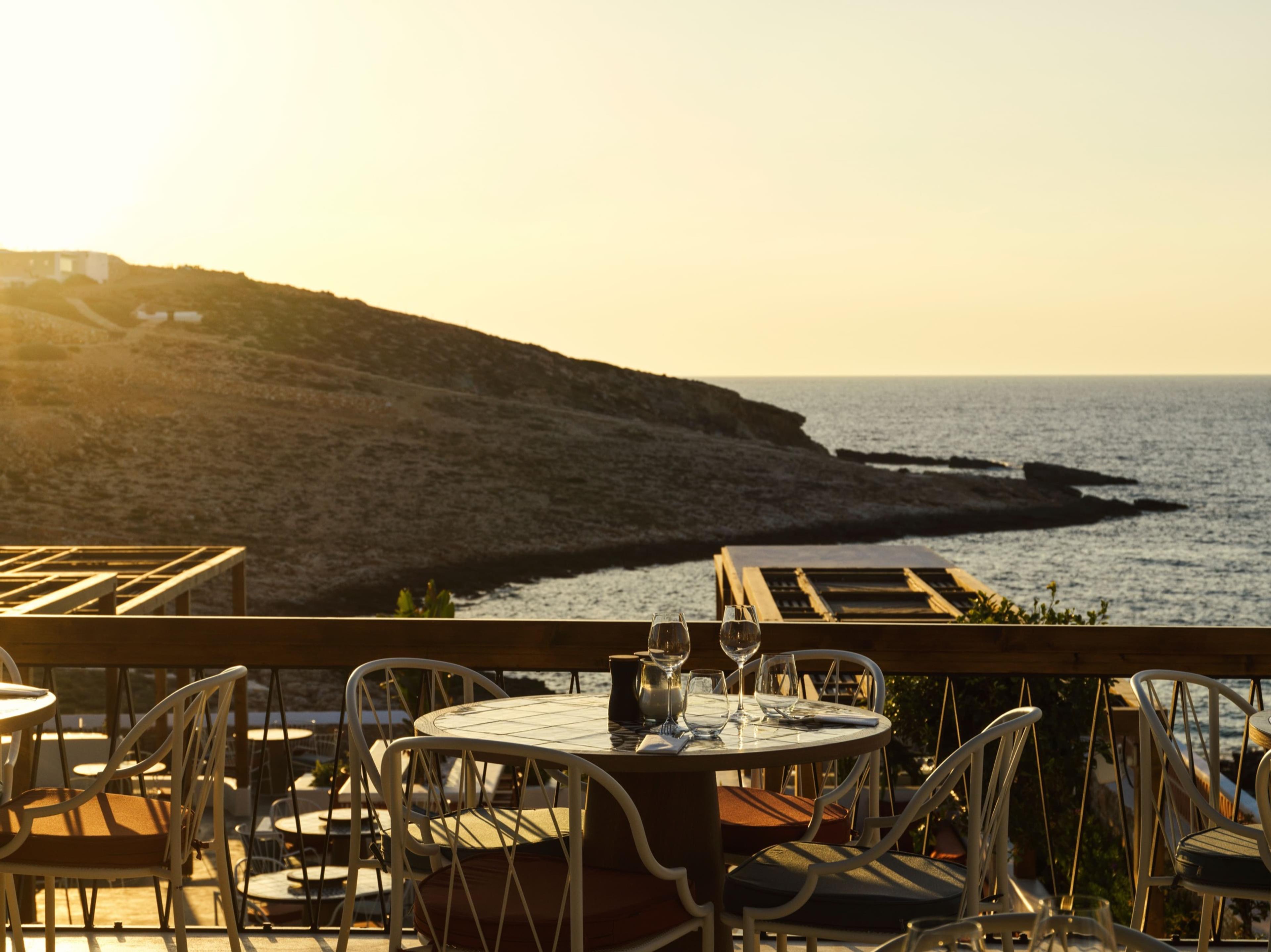 A round table set with wine glasses stands on an outdoor terrace at sunset, with chairs around it and a rocky headland and the sea beyond the railing.