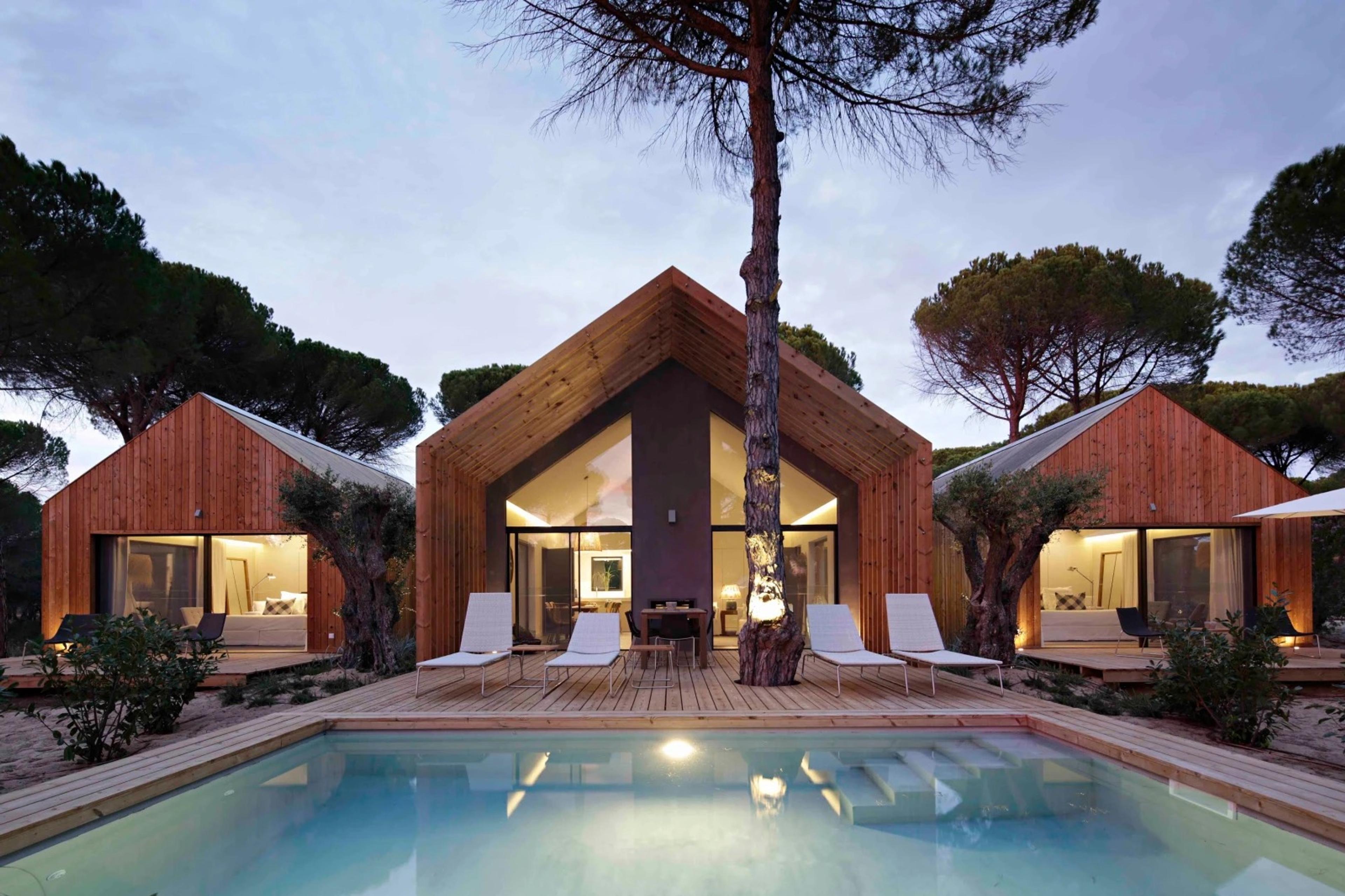 Three wood-clad cabins lit at dusk around a pool, with a tall pine tree rising through the deck between them.