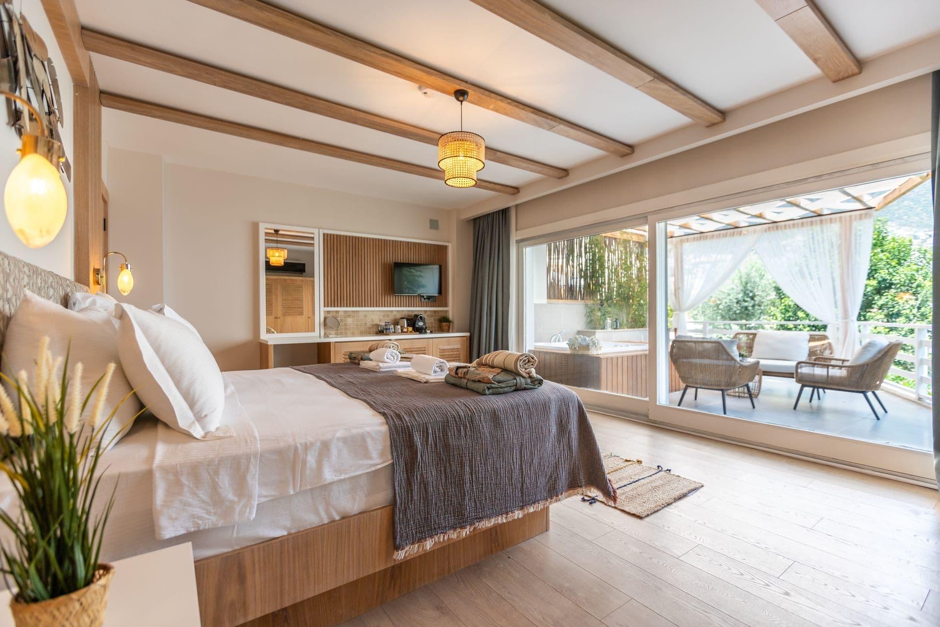 A bed with a dark throw sits opposite glass doors that open onto a covered terrace with wicker chairs and a sunken tub.