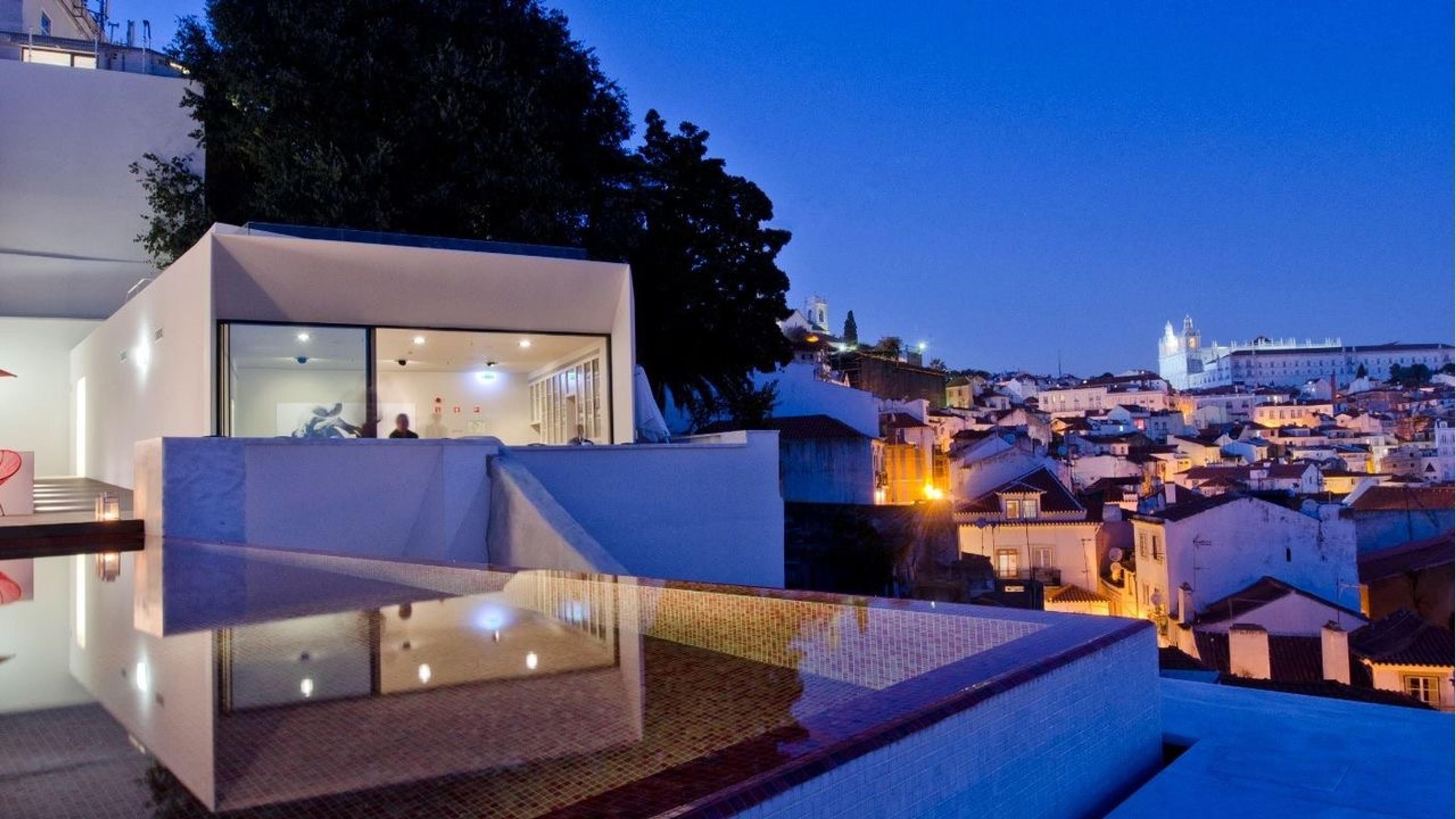 A low white building with a glass-walled room lit from inside, beside a pool with a tiled edge, with red-roofed houses on a hillside below a church with two towers, all after dark.