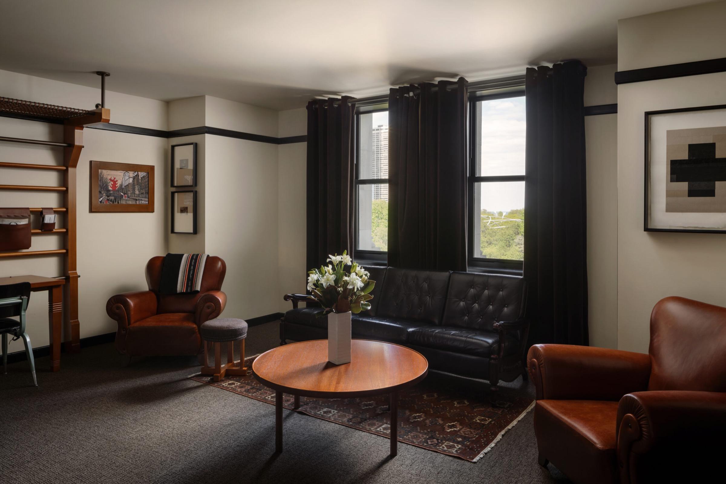 A sitting area with a black leather sofa, two leather armchairs, and a round wood coffee table, in front of tall curtained windows looking onto a city skyline and trees.