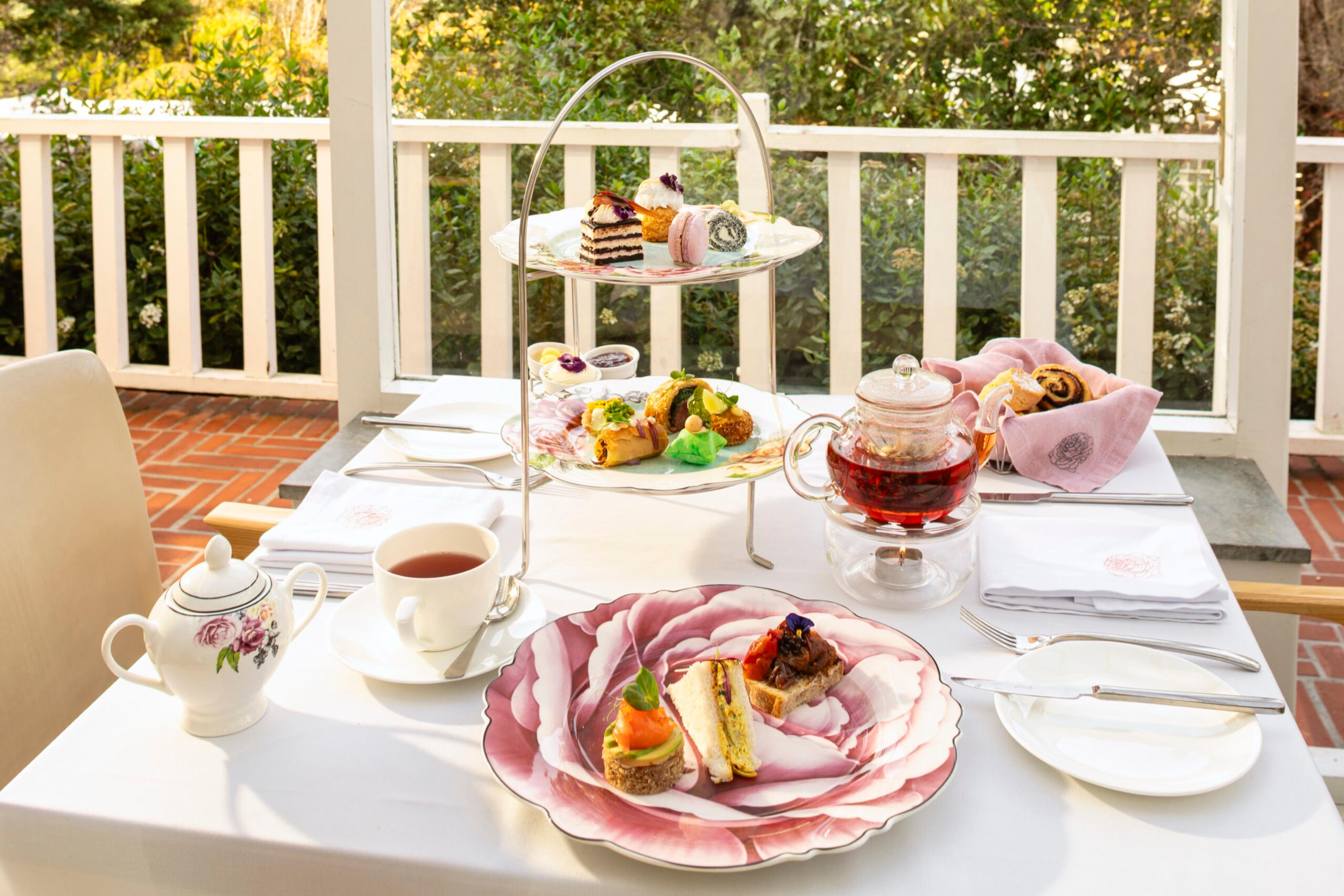 Afternoon tea on a white table on a veranda, with a tiered stand of cakes, a teapot, and sandwiches on a pink-rimmed plate.