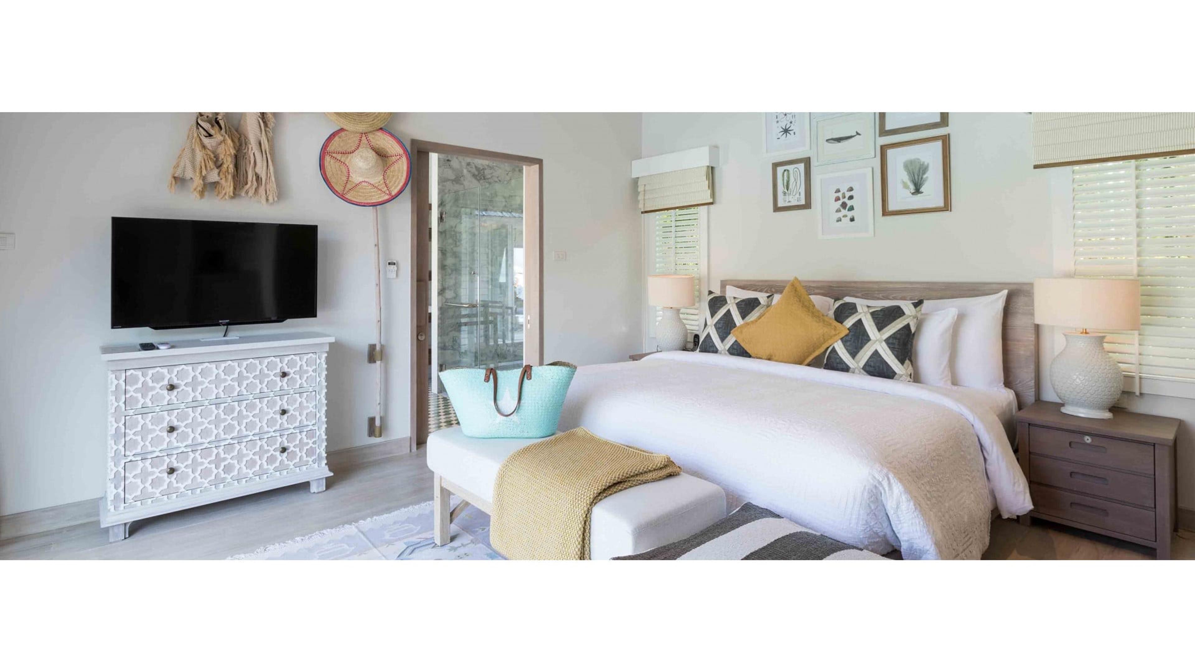 A bedroom with a white bed dressed in mustard and grey pillows, a dresser holding a television, and framed botanical prints on the wall.