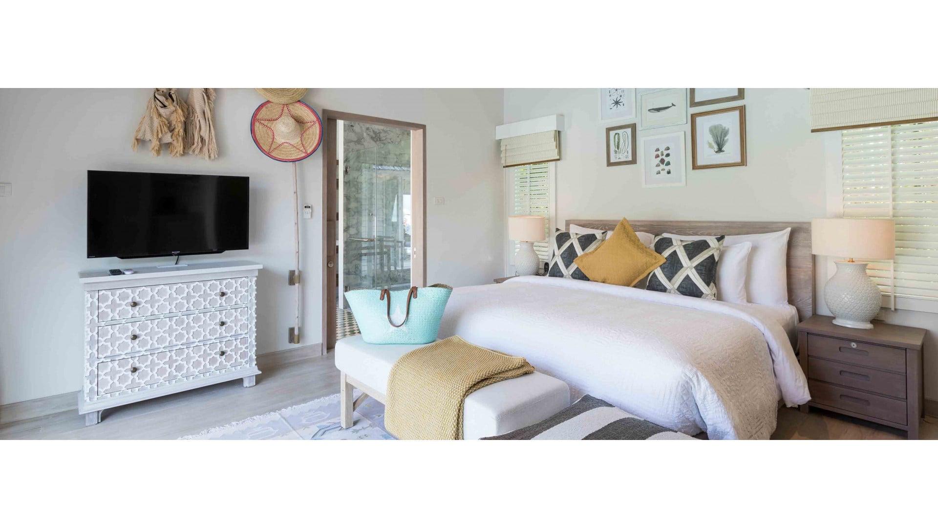 A bedroom with a white bed dressed in mustard and grey pillows, a dresser holding a television, and framed botanical prints on the wall.