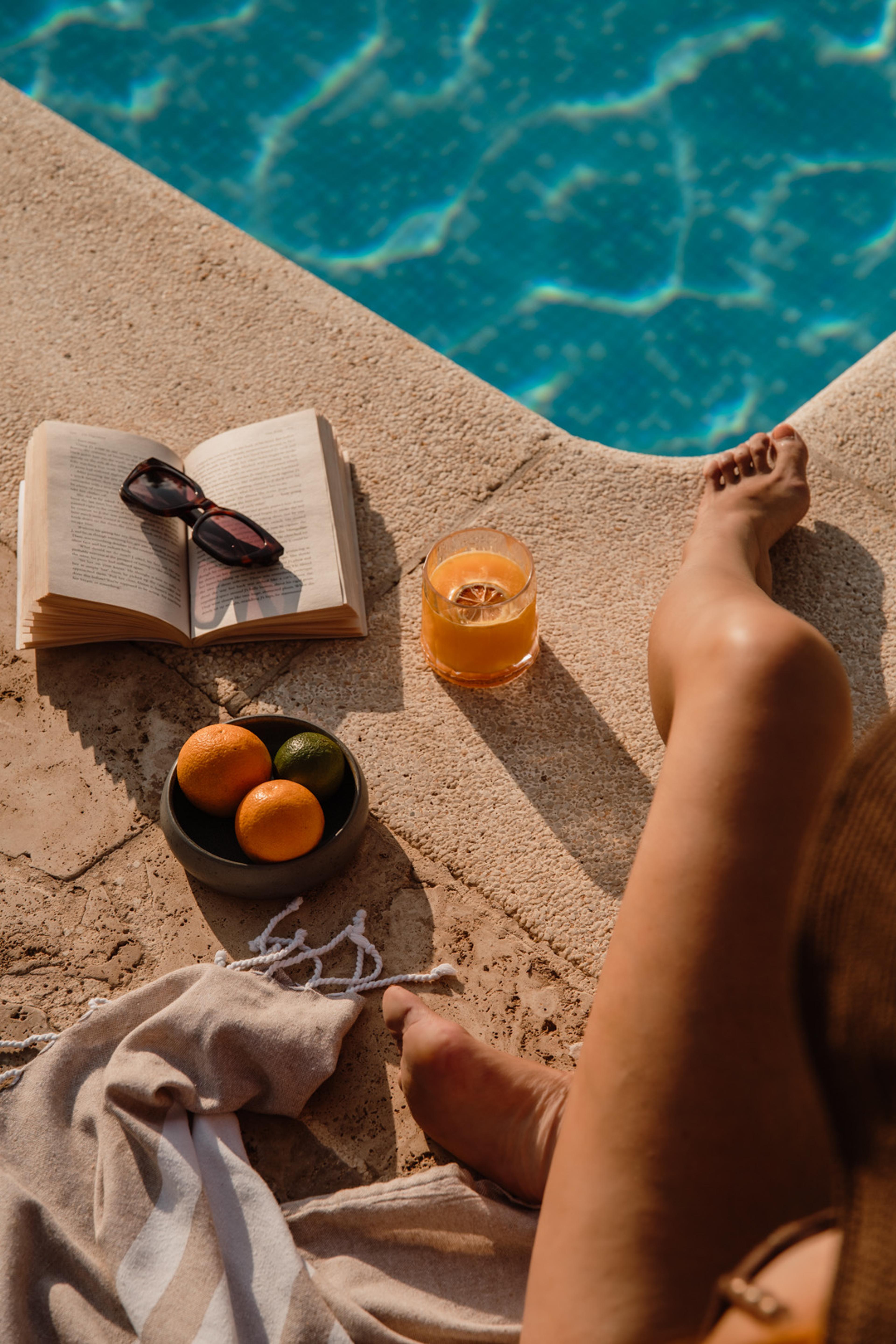 A view looking down at an open book, sunglasses, a bowl of oranges and a glass of juice on a pool ledge, with bare feet and a folded towel nearby.
