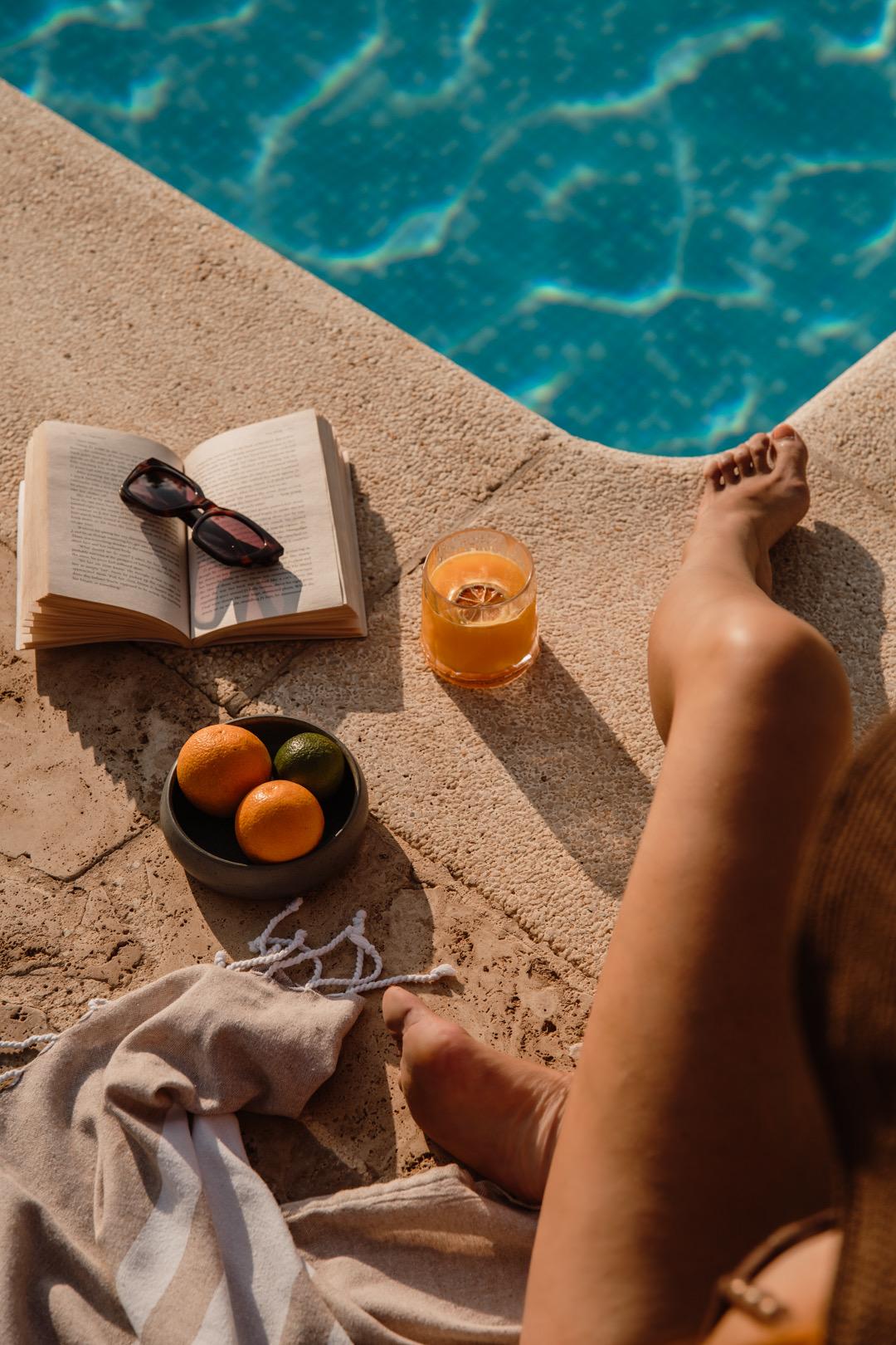 A view looking down at an open book, sunglasses, a bowl of oranges and a glass of juice on a pool ledge, with bare feet and a folded towel nearby.
