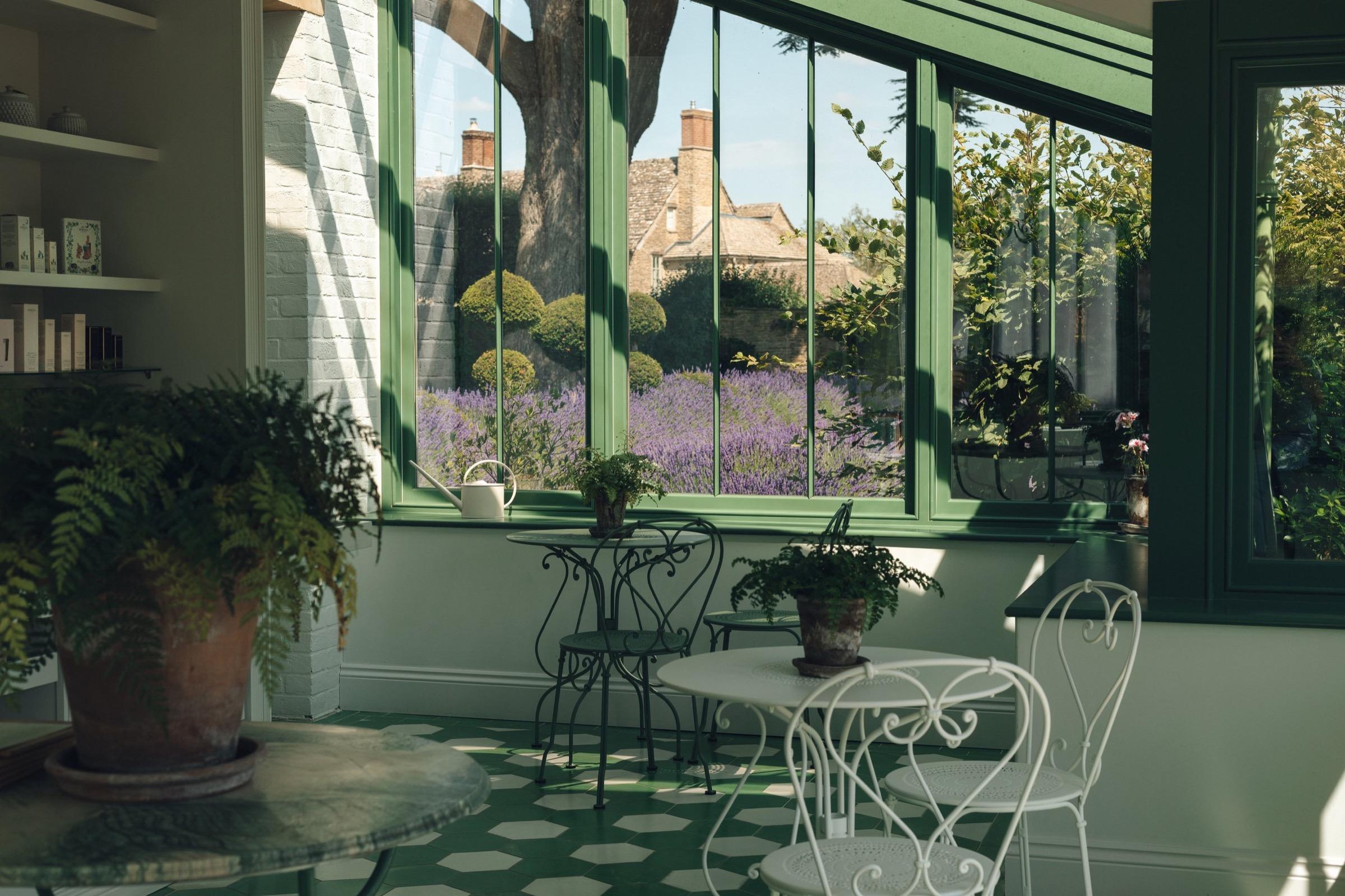 A conservatory with wicker cafe chairs and tables around potted ferns, its windows open to a lavender border and a stone house beyond.