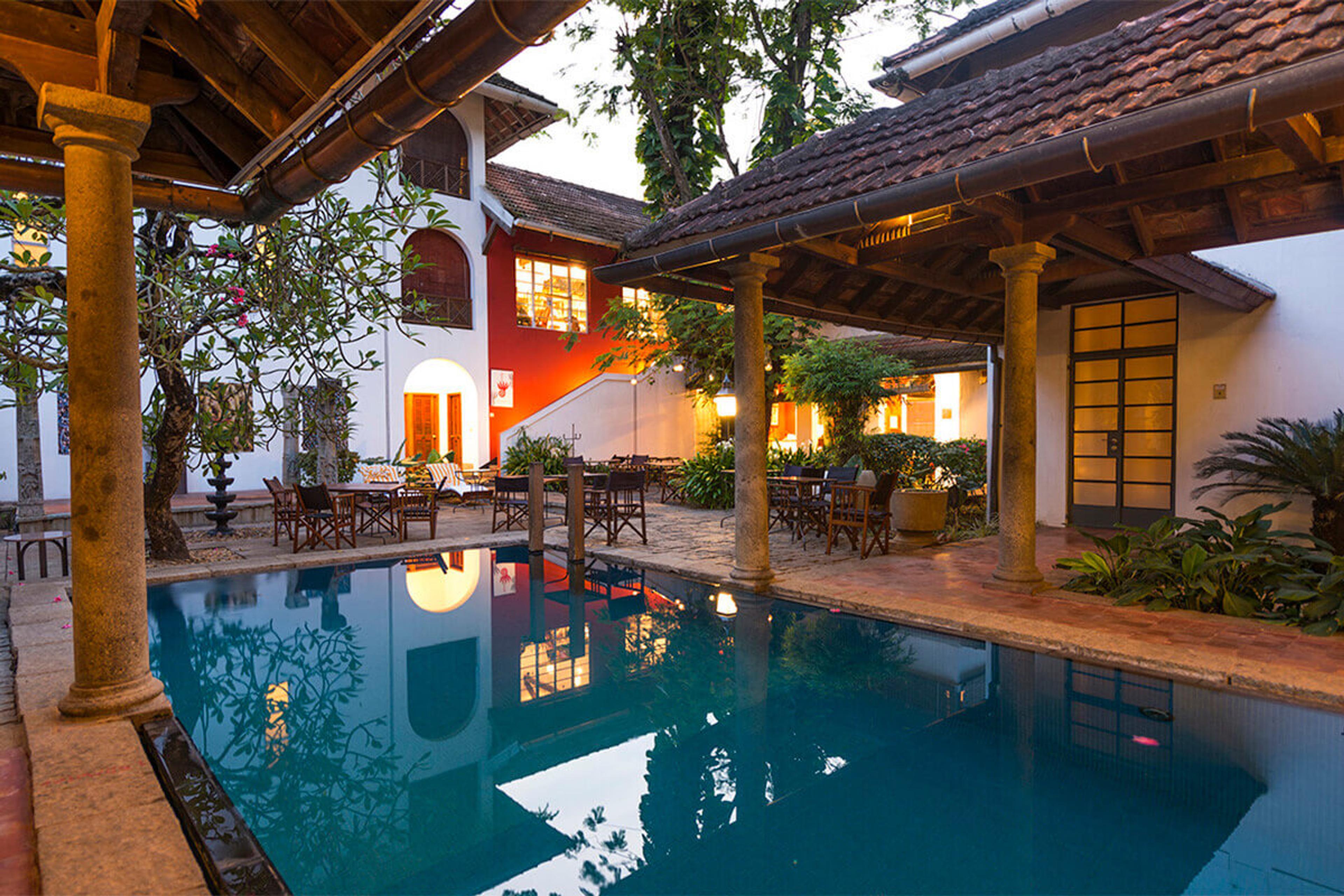 A lit swimming pool at dusk beside a tiled-roof courtyard building, with dining tables set out under the eaves and the lit windows reflected in the water.