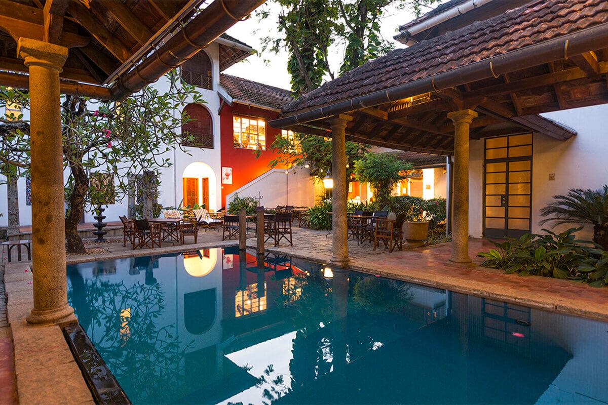 A lit swimming pool at dusk beside a tiled-roof courtyard building, with dining tables set out under the eaves and the lit windows reflected in the water.