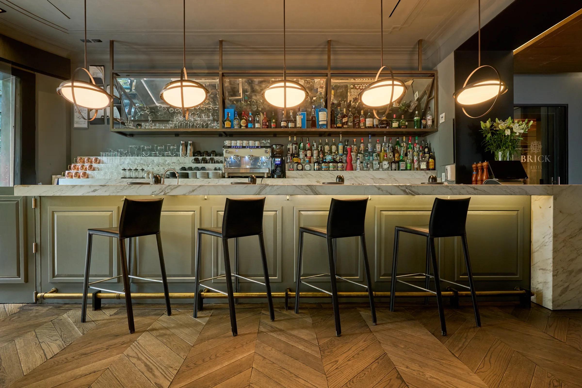 A hotel bar with four black stools at a marble counter, bottles and glasses on shelves behind, and round pendant lights overhead.