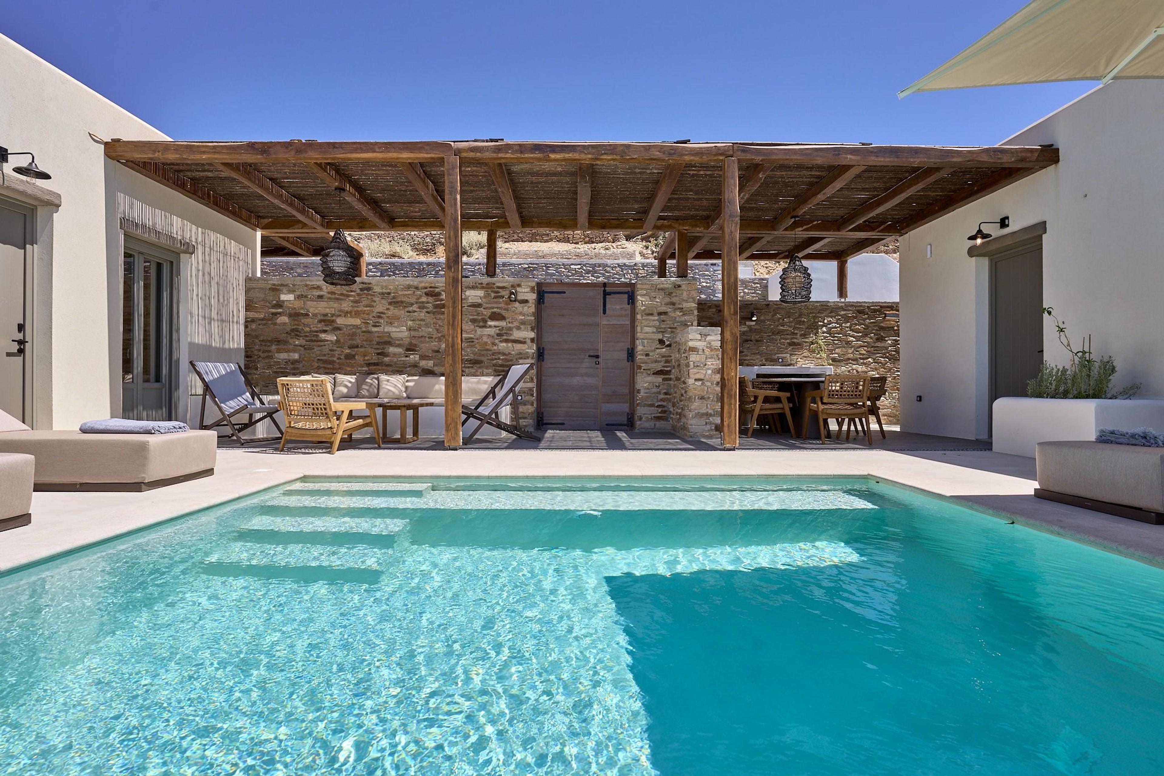 A small courtyard pool beside a sofa and a dining table under a timber pergola, with stone walls and a wooden door behind.