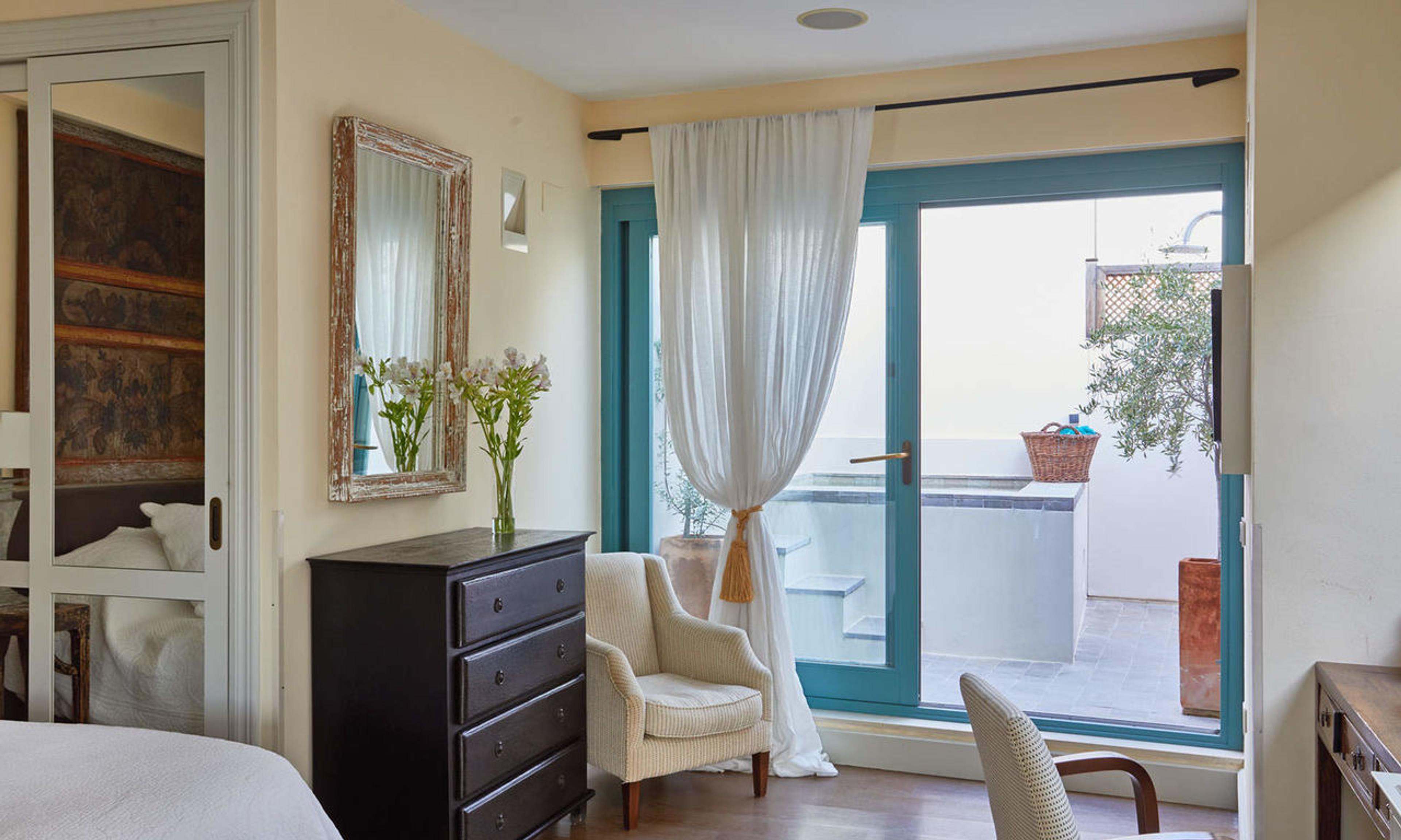 A bedroom with a dark chest of drawers, an upholstered armchair and a curtained glass door open onto a tiled terrace with a plunge pool and terracotta pots.